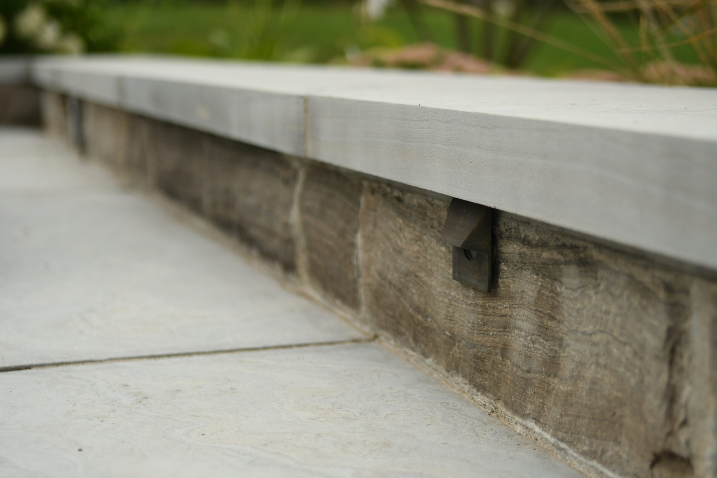 Close-up of stone and concrete ledge with metal piece, surrounded by greenery in the background, featuring a minimalist outdoor setting.