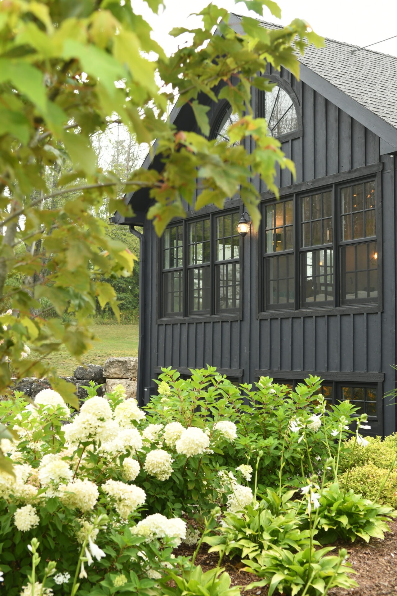 A charming black cottage with large windows, surrounded by lush greenery and white hydrangeas, creates a serene, picturesque garden setting.