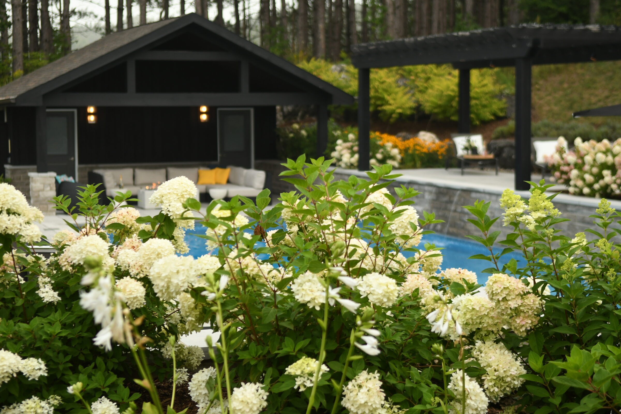 Lush garden with white flowers, a swimming pool, and cozy seating area under a dark pavilion; serene and inviting outdoor space.