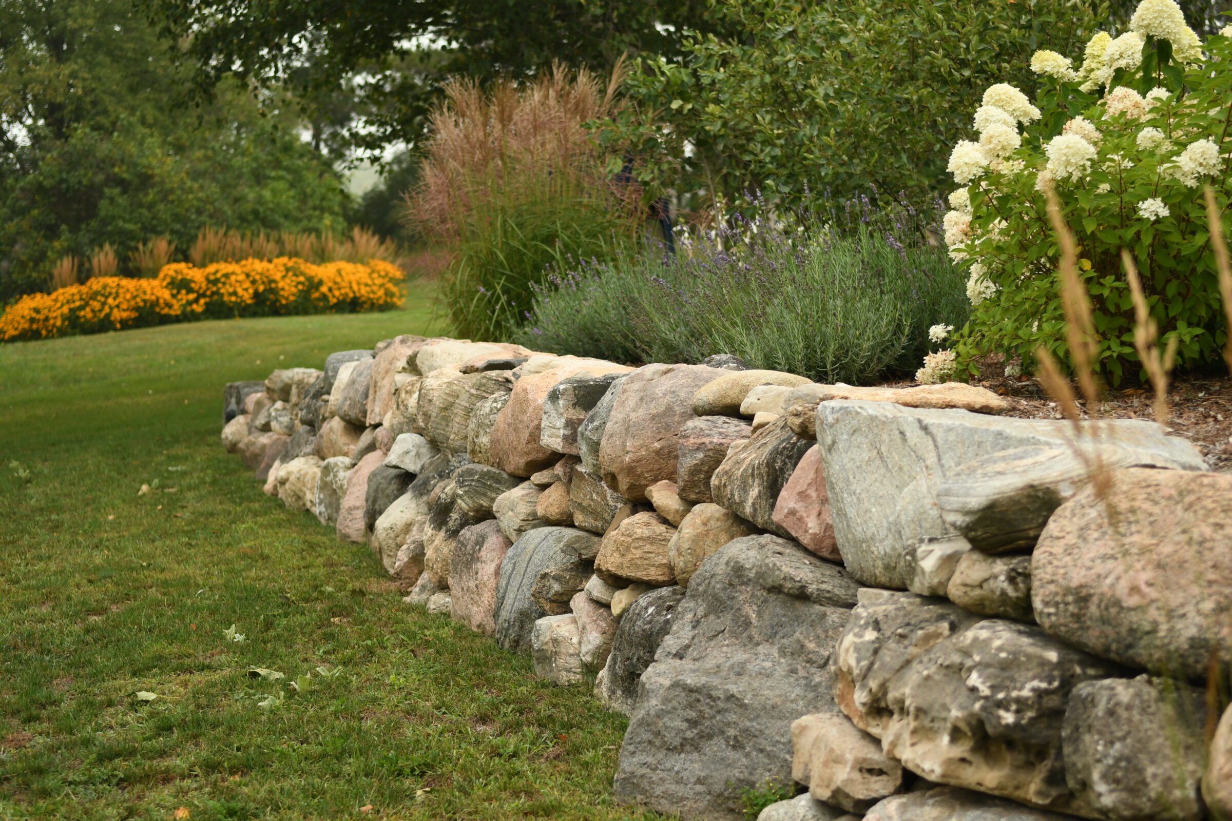 A stone wall borders a lush garden with vibrant flowers and greenery, creating a serene, landscaped setting under overcast skies.