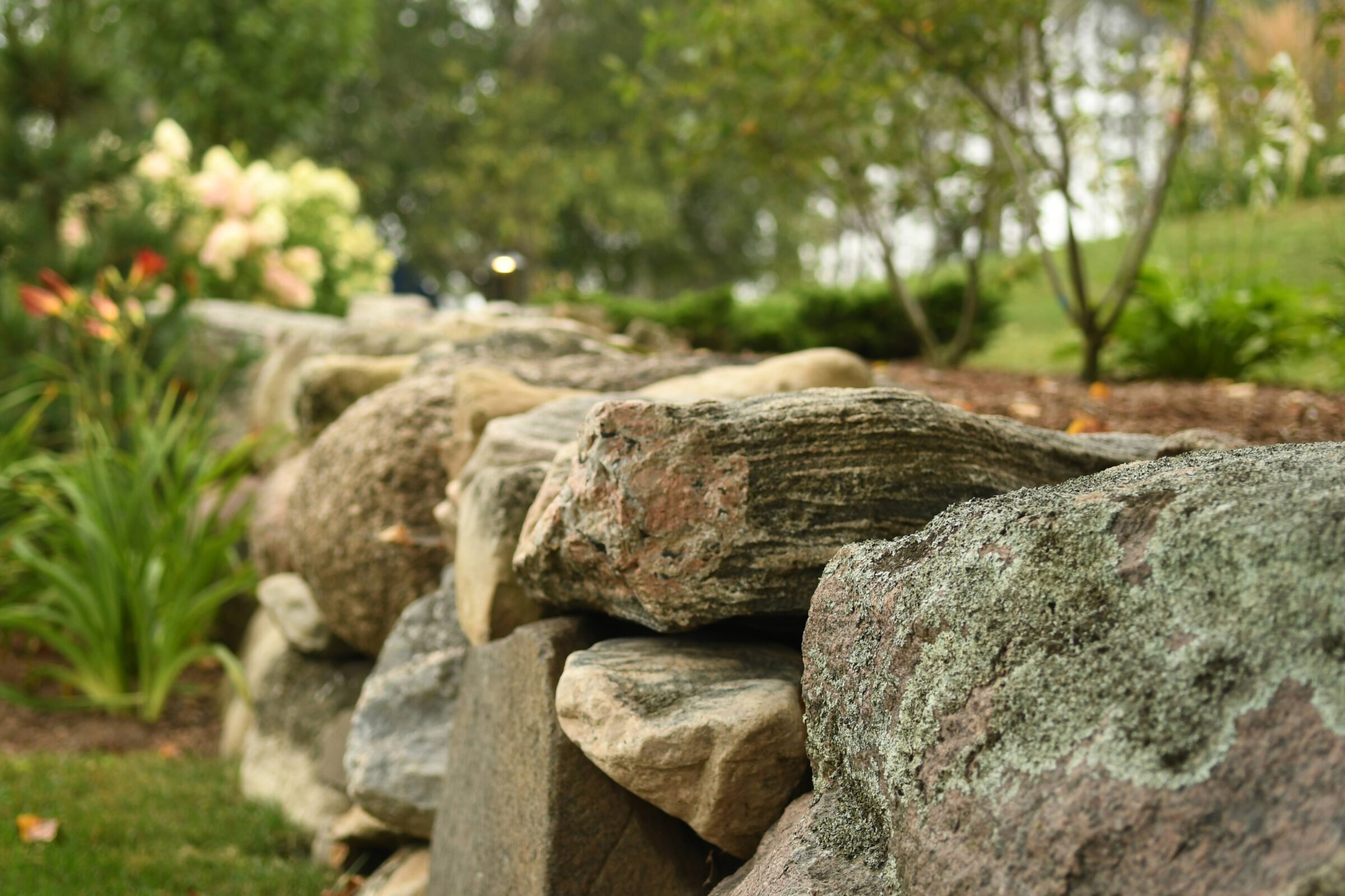 A close-up of a stone wall in a lush garden, surrounded by greenery and flowers, with soft background focus.