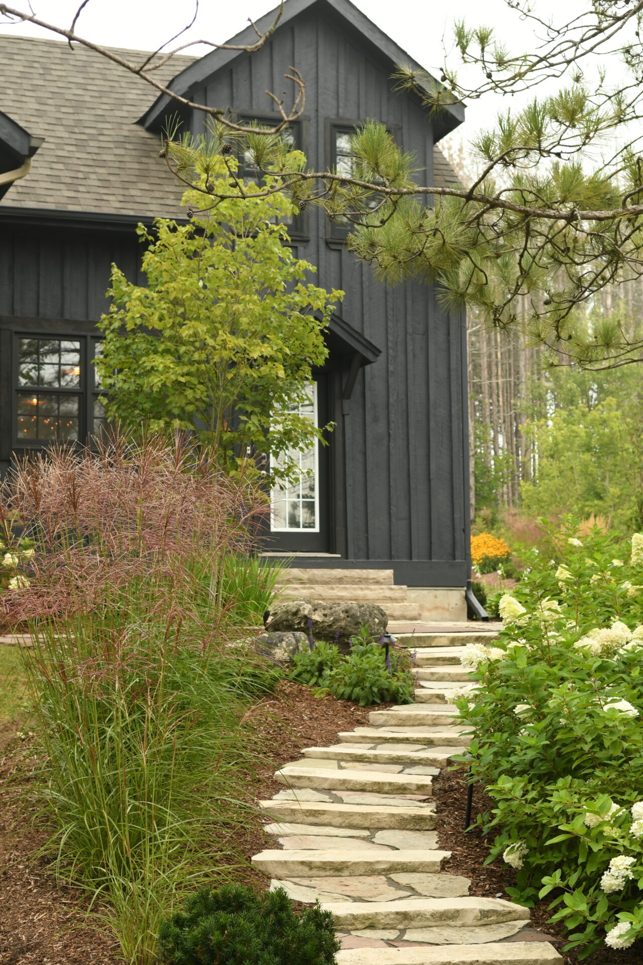 A cozy black house with a stone pathway is surrounded by lush greenery, featuring trees and flowering plants in a peaceful garden.