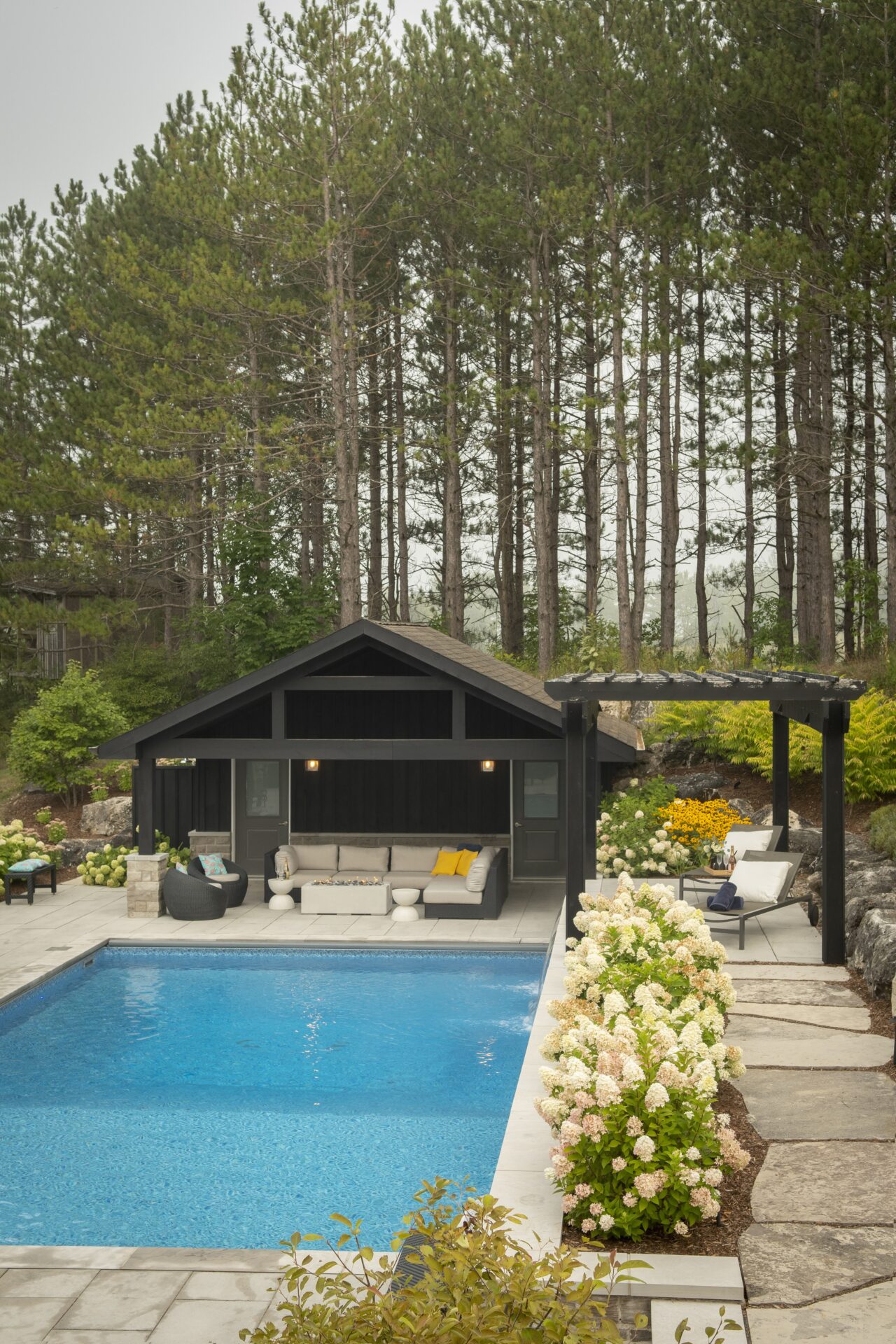 A poolside retreat features a cozy seating area, surrounded by lush greenery and vibrant flowers, situated beneath tall, dense pine trees.