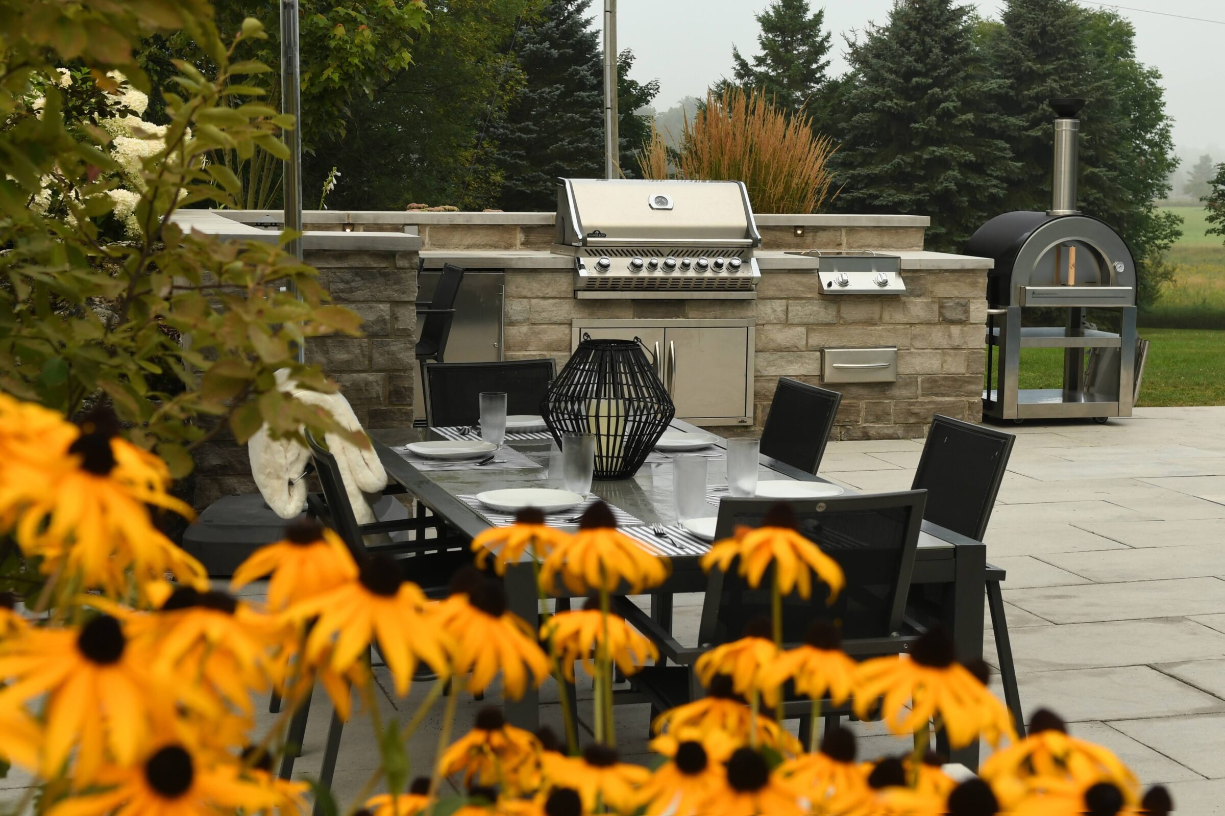 Outdoor kitchen with grill, dining table, and chairs surrounded by yellow flowers and lush greenery on a patio.