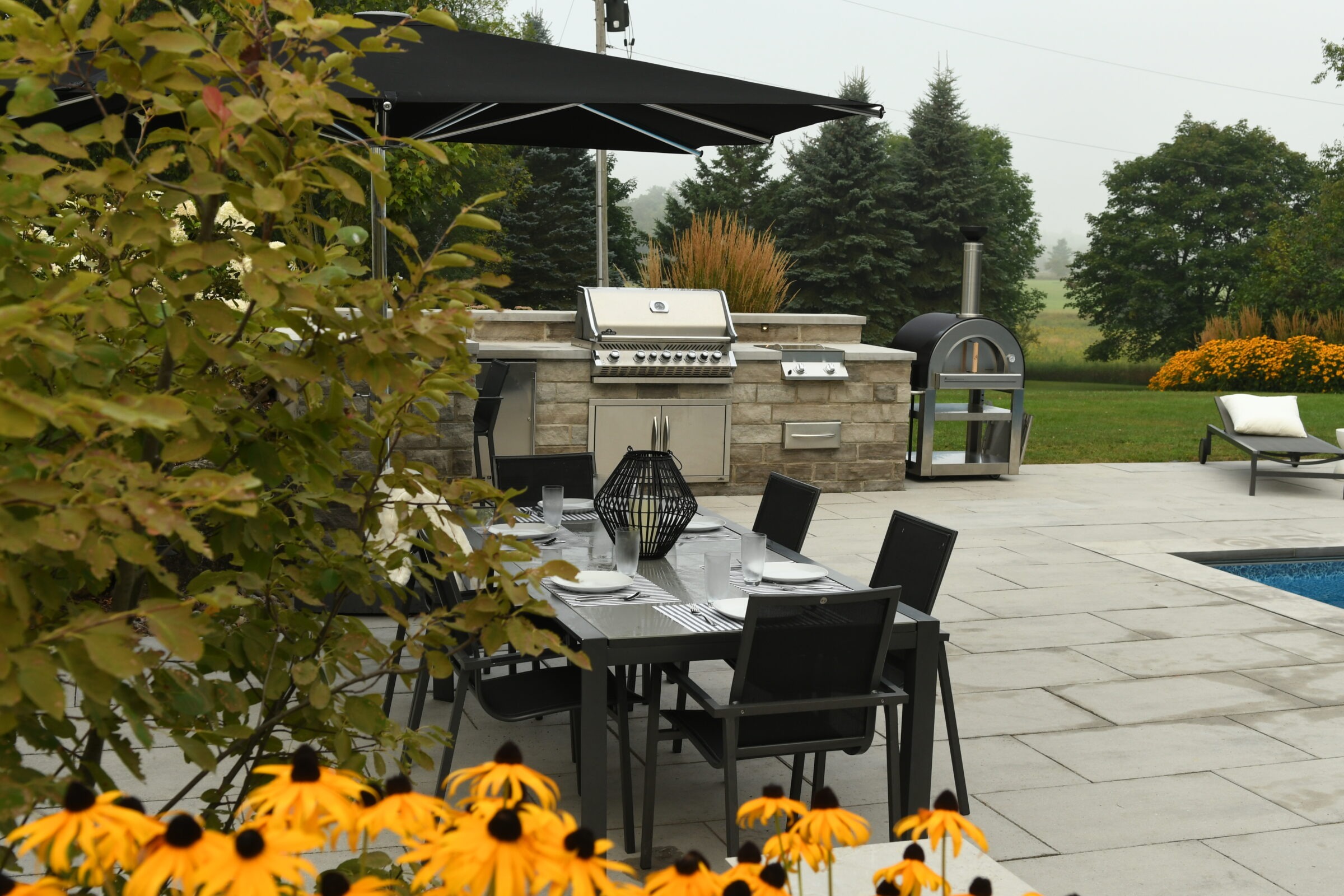 Outdoor patio with dining table, black furniture, grill, and pool. Surrounding greenery includes yellow flowers and trees, creating a serene garden setting.