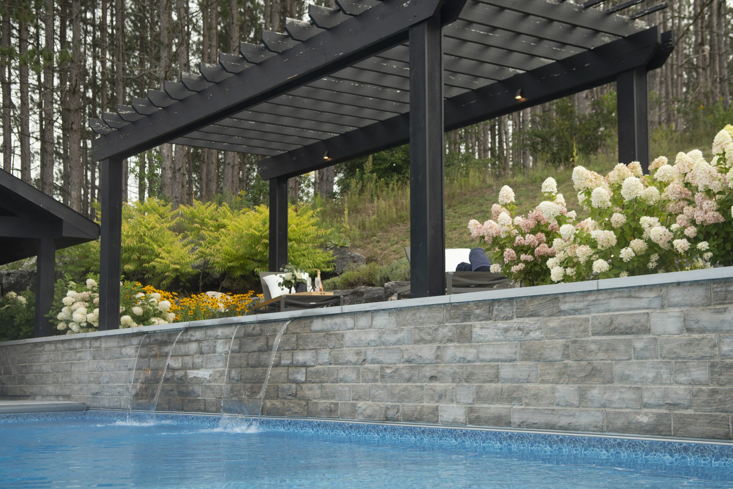 A peaceful poolside scene with a pergola, blooming flowers, and a person relaxing on a lounge chair amidst a wooded backdrop.