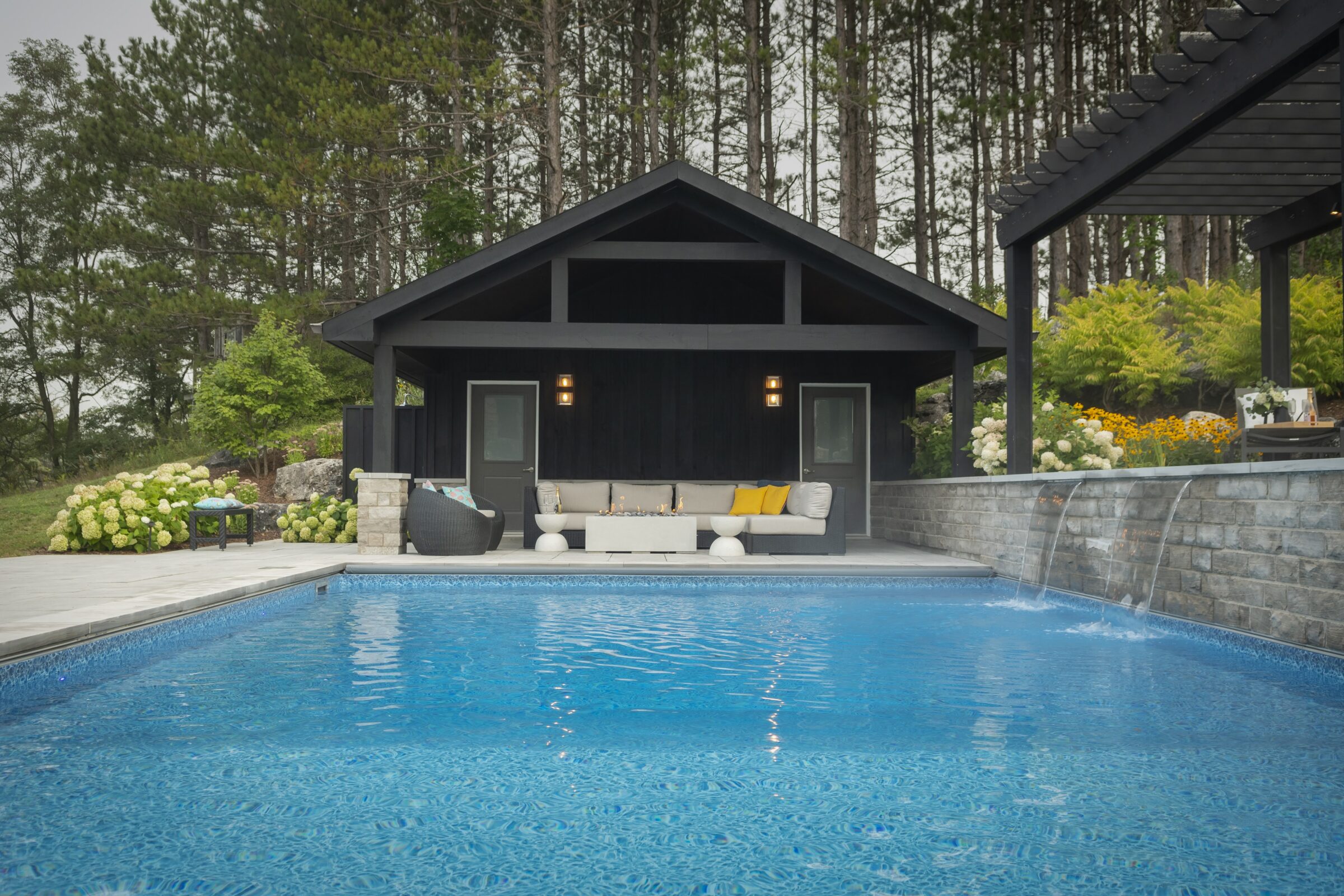 A serene poolside view features a cozy cabana with comfortable seating, surrounded by lush trees and vibrant flowers, creating a tranquil retreat.