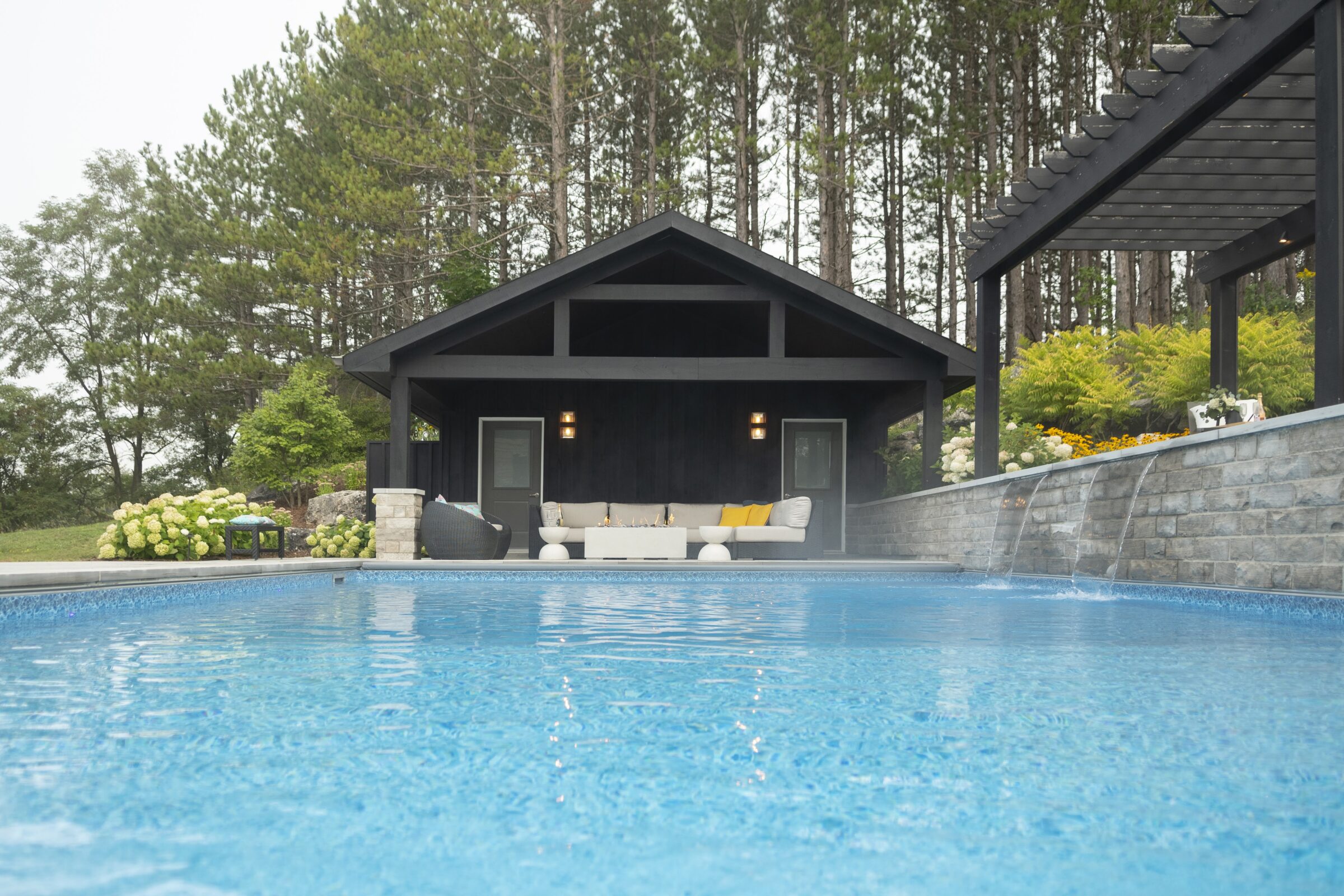 A serene backyard scene features a clear pool, modern pavilion, lounge area, and lush greenery against a backdrop of tall trees.