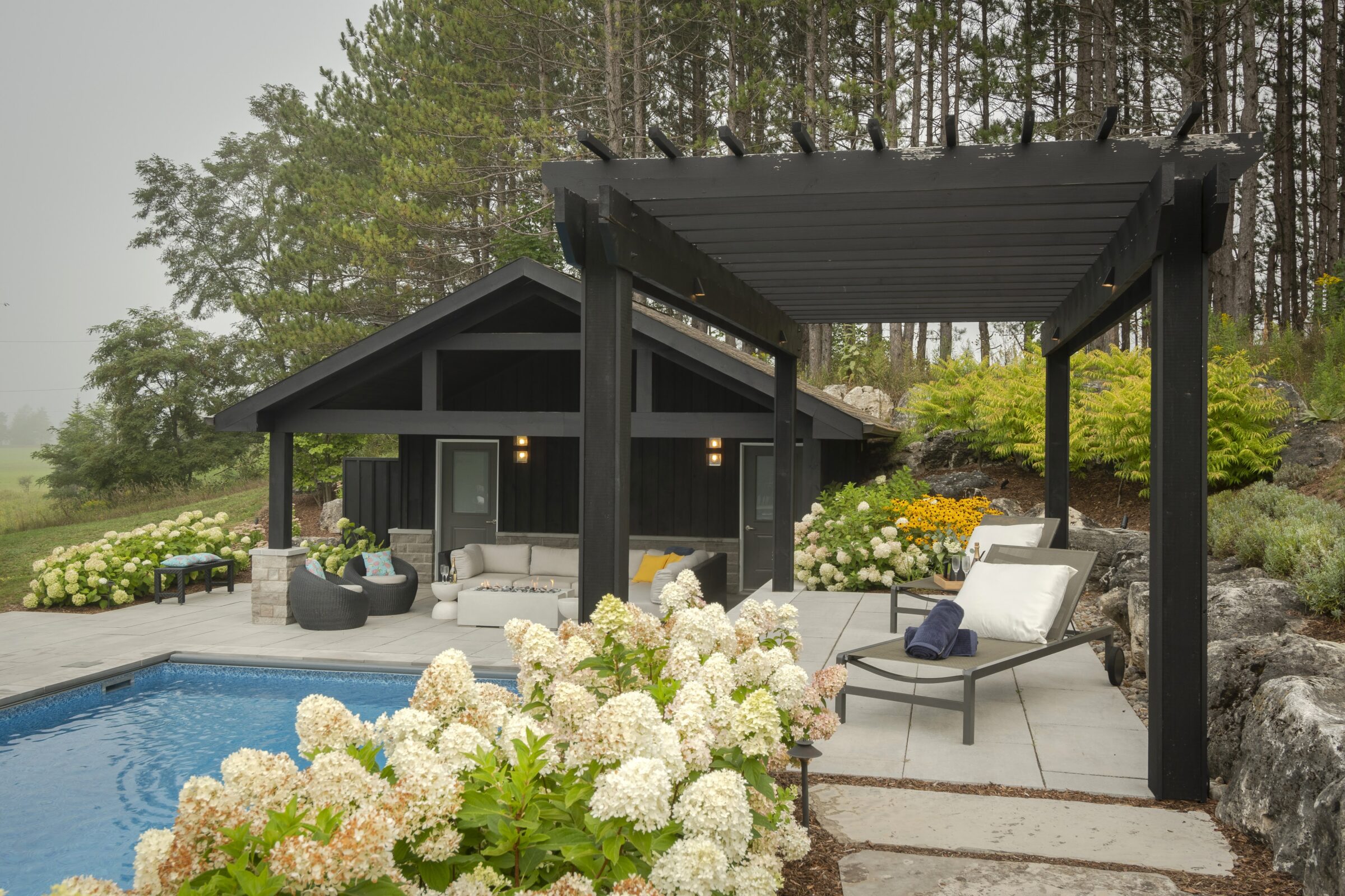 A serene backyard features a modern pergola, cozy seating, lush greenery, and a pool, surrounded by trees and flowering plants.