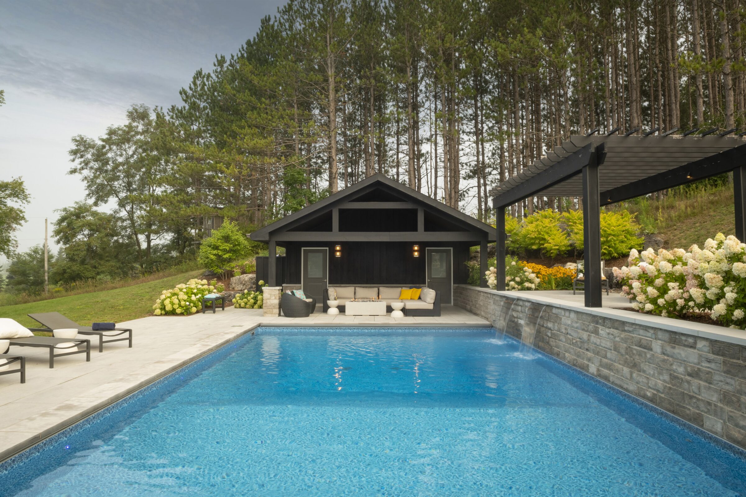 A serene backyard features a bright blue swimming pool, bordered by a modern pool house and lush garden, surrounded by tall trees.
