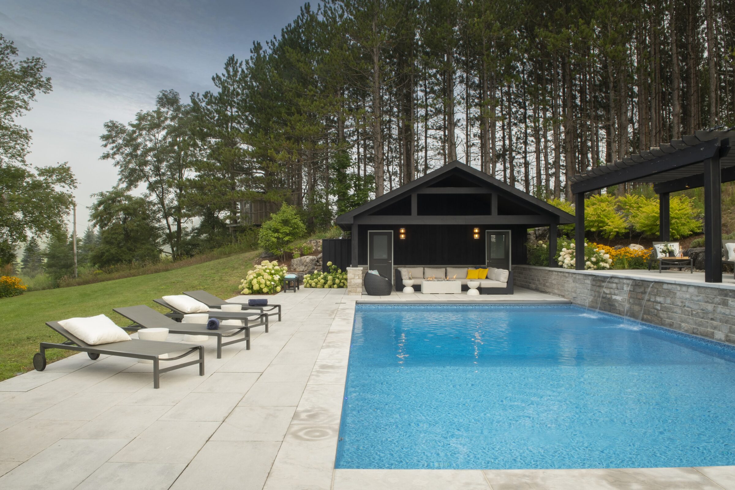 Modern backyard with a pool, several lounge chairs, a patio area, surrounded by greenery and trees, next to a cozy pool house.