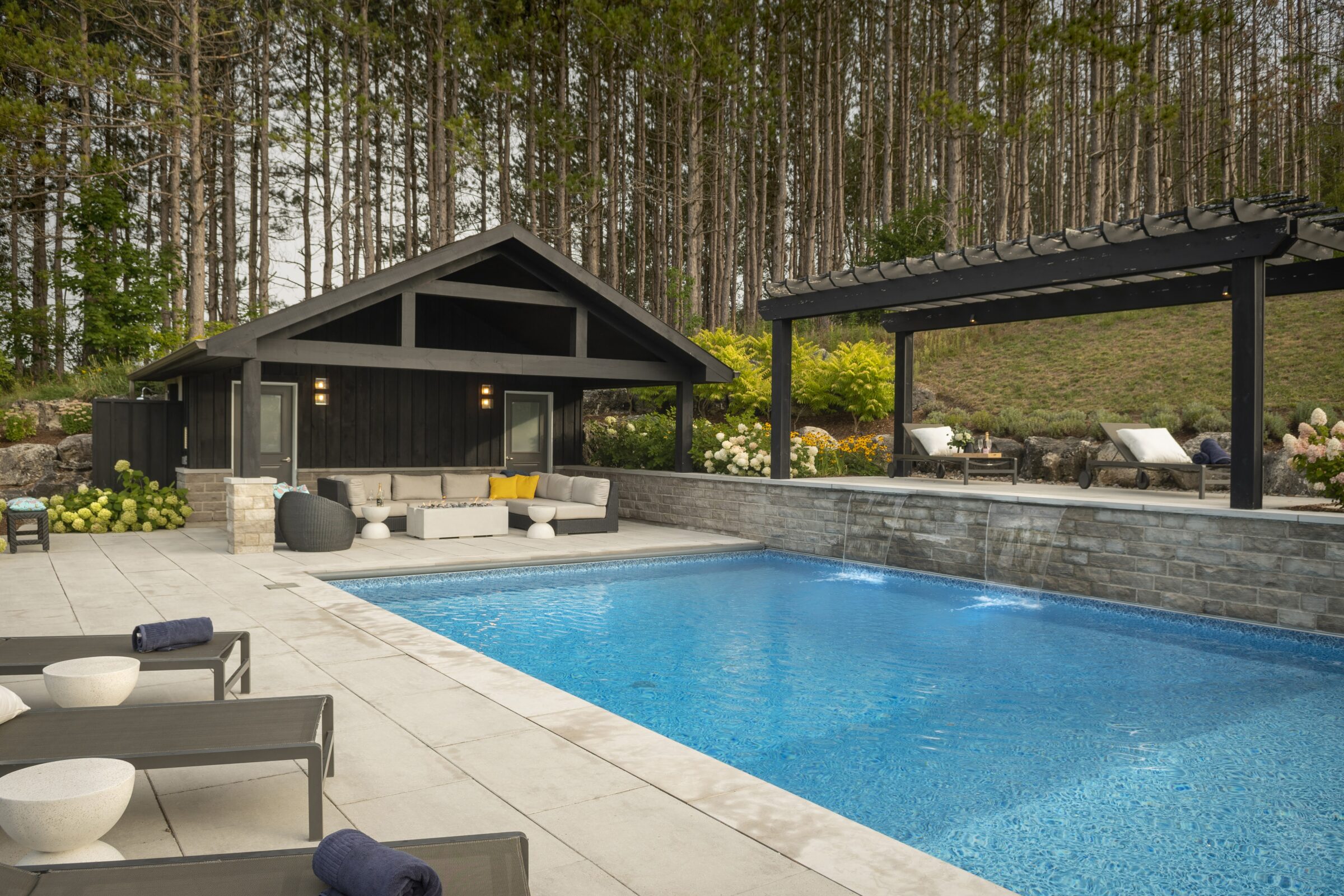 Modern outdoor pool area with cozy seating, loungers, and a pergola surrounded by lush greenery and tall trees, creating a serene retreat.