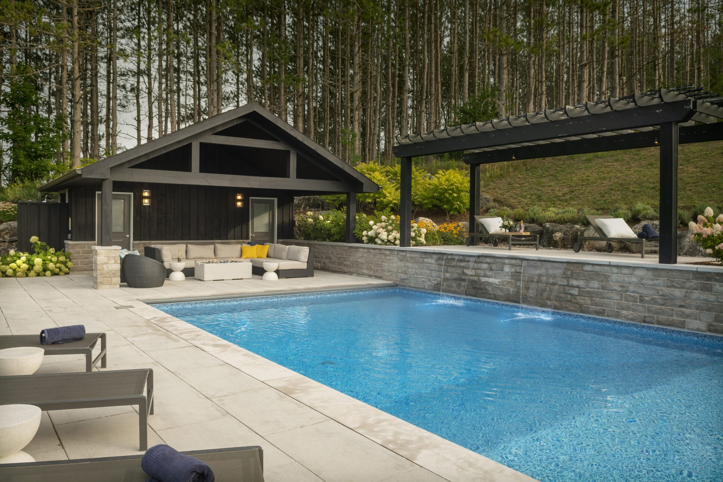 Modern outdoor pool with stone patio, lounge chairs, and pergola. Cabin-like structure in background, surrounded by pine trees and lush greenery.