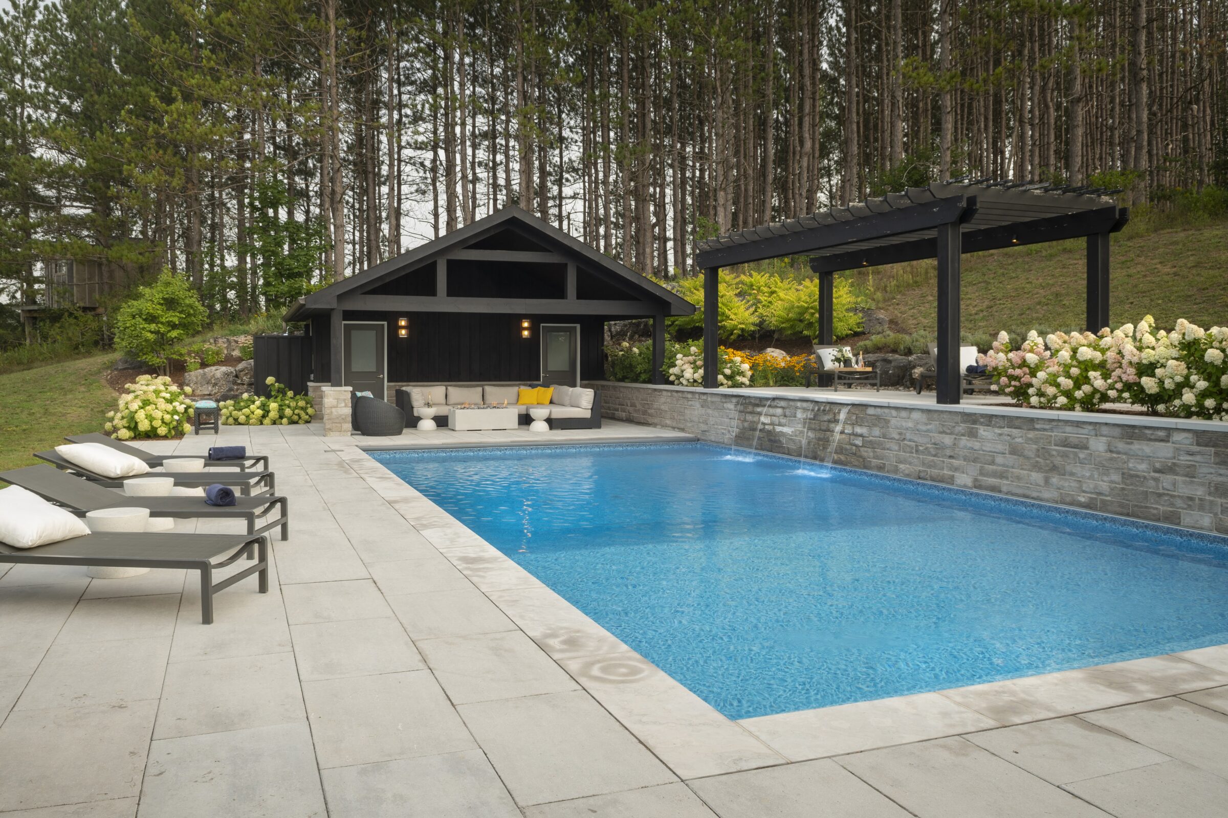 Backyard features a modern pool, lounge chairs, and shaded seating area surrounded by lush landscaping and a small black pool house amidst tall trees.