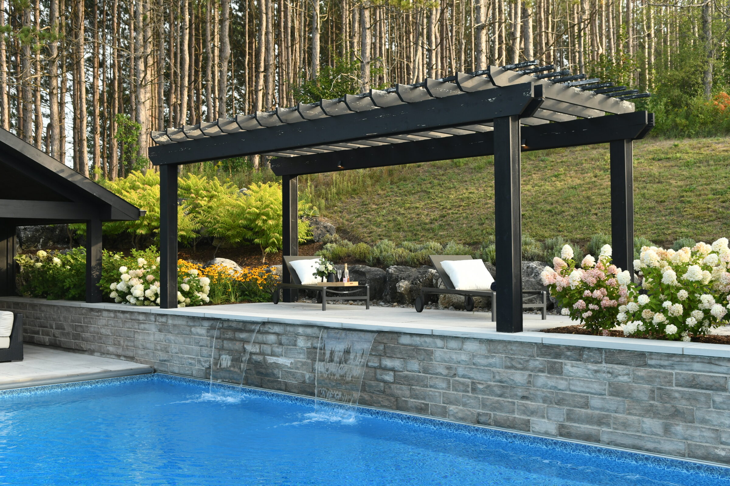 A serene backyard features a modern pergola, blooming hydrangeas, and a pristine pool with waterfalls, surrounded by a lush forest.