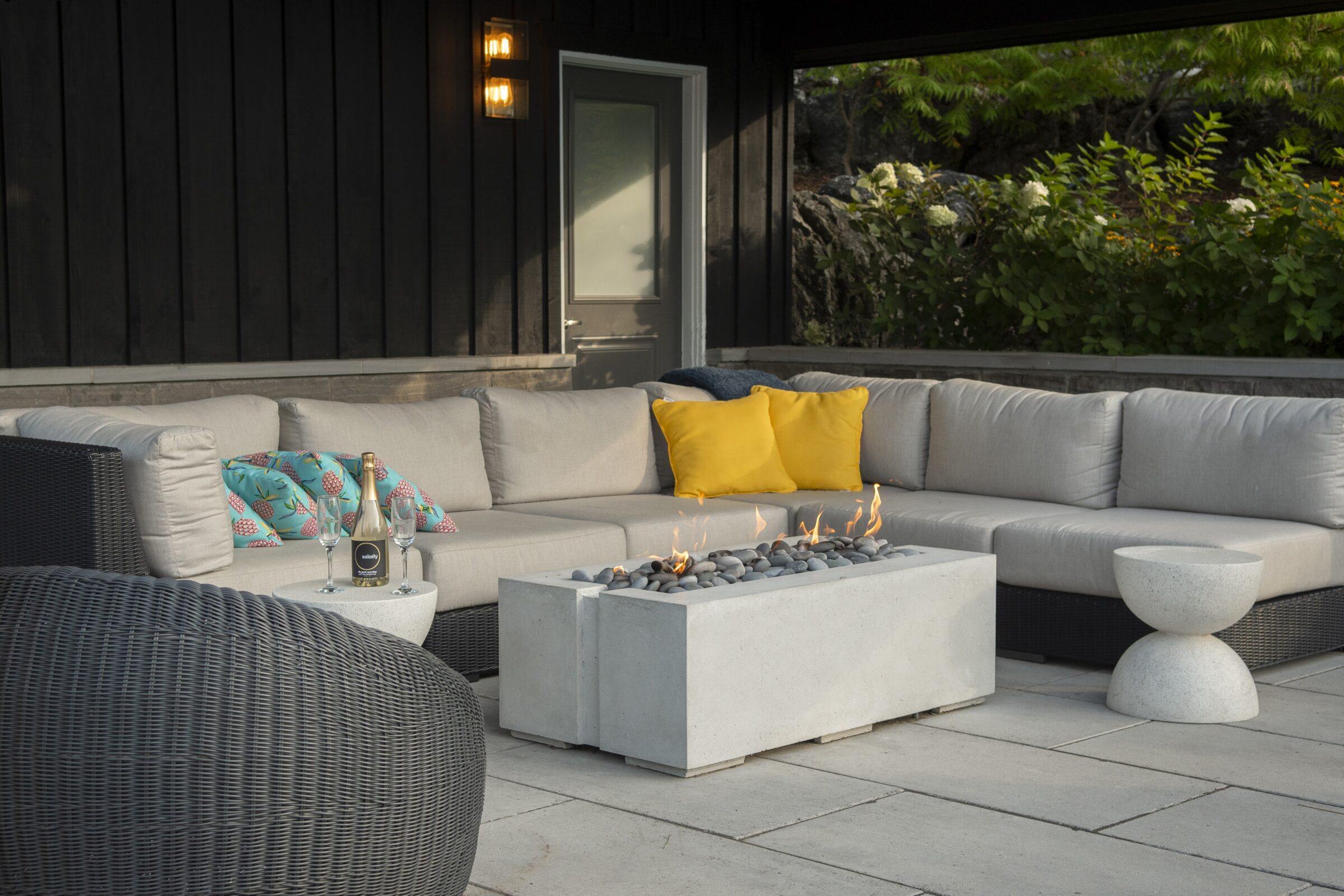 Outdoor patio with gray sectional sofa, yellow pillows, a gas fire pit, and drinks set on a round table. Surrounded by greenery.