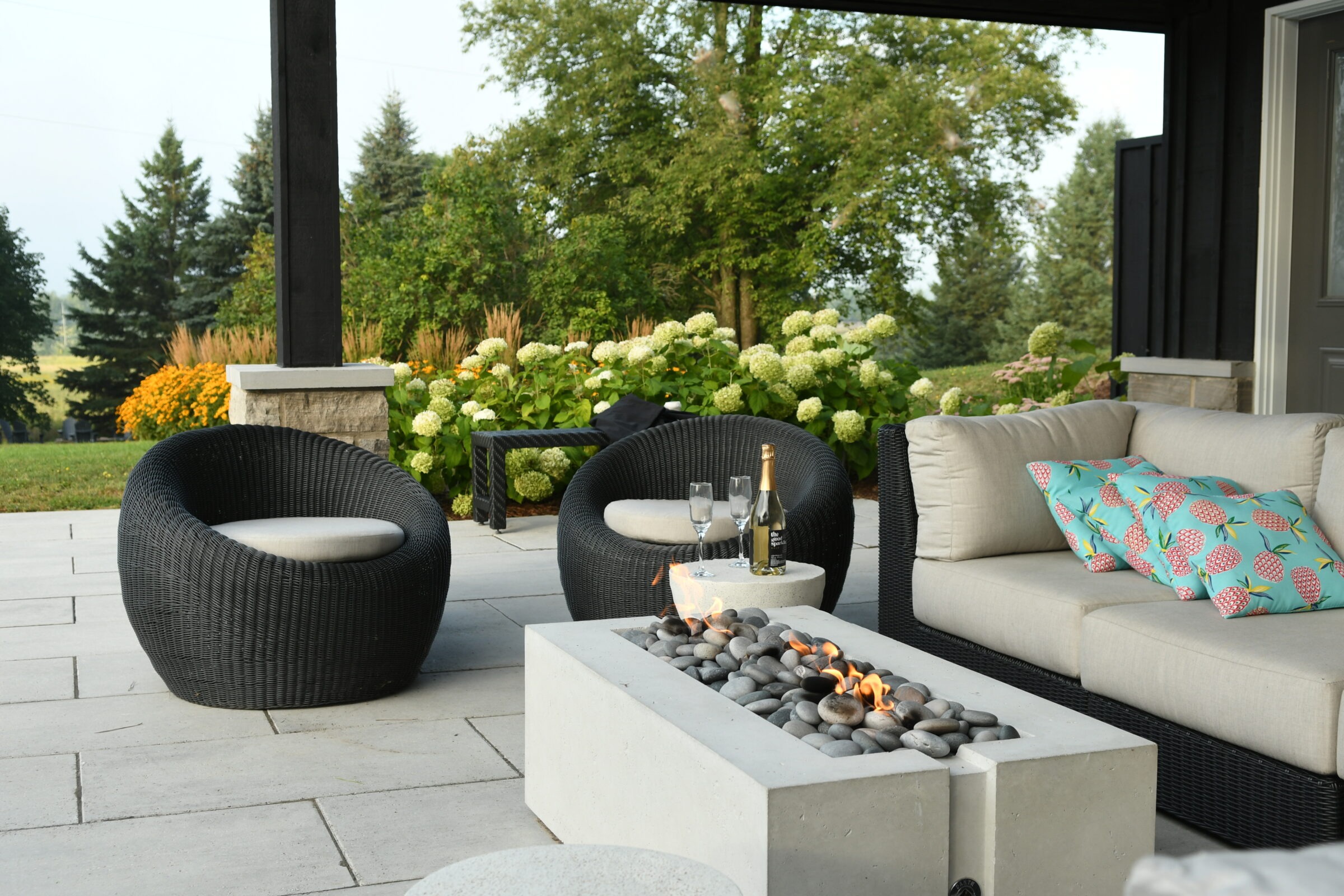 Modern patio with black chairs, cushioned sofa, and a rectangular fire pit. Champagne and glasses are on the table. Lush greenery surrounds.