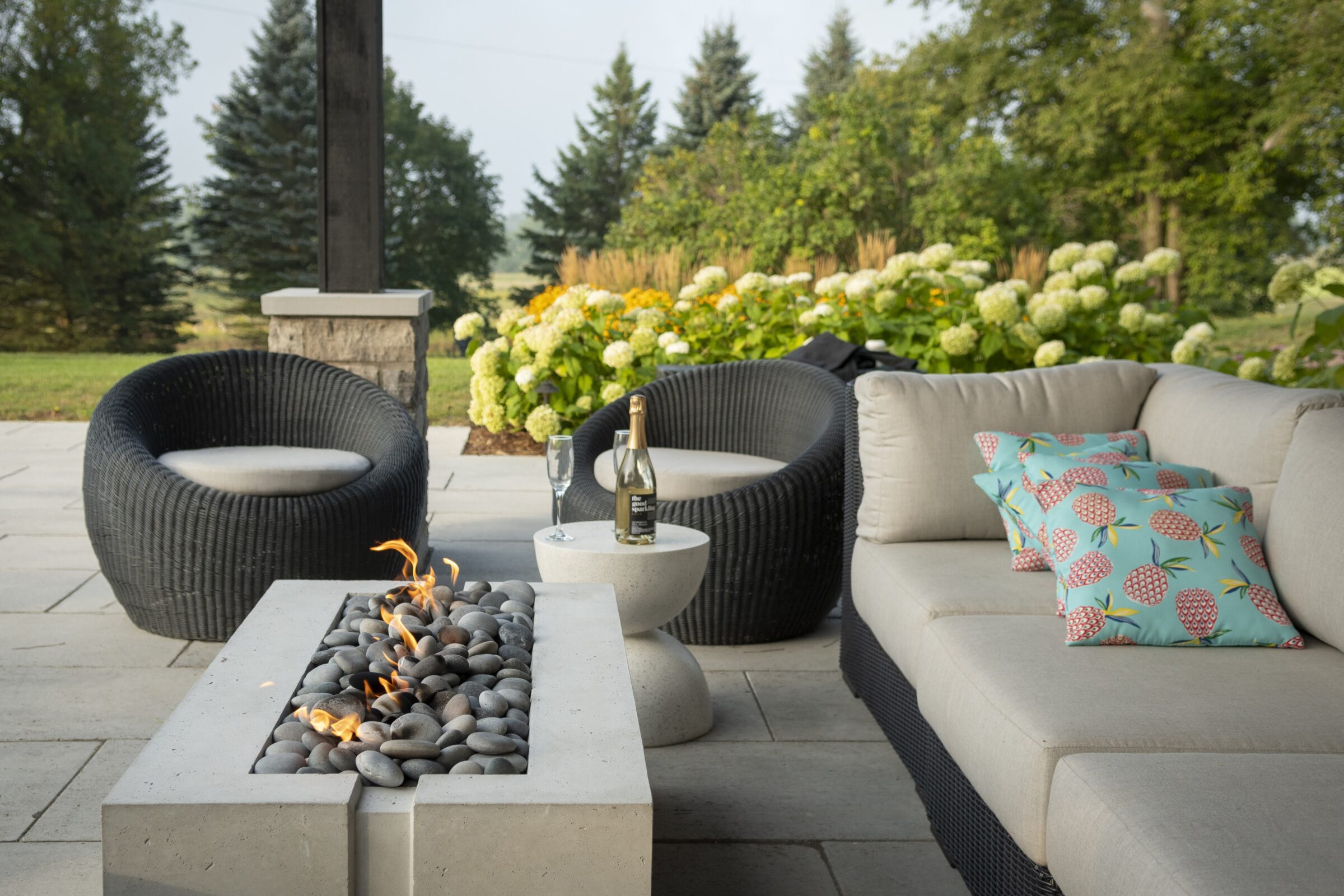 Cozy outdoor patio with modern furniture, fire pit, and garden view. Bottled drink and glasses on small table, surrounded by greenery.