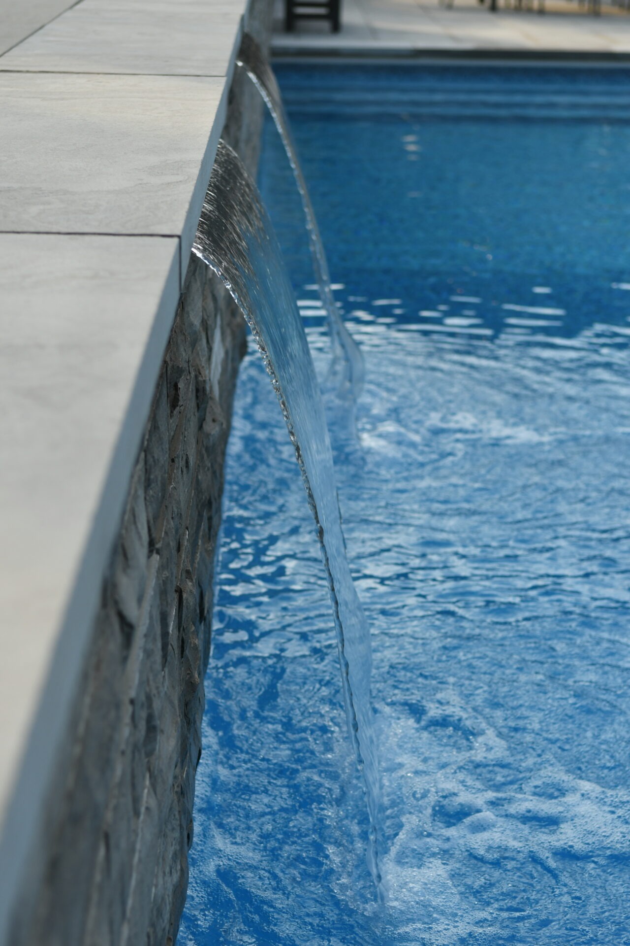 A serene poolside scene with water cascading over a stone wall into a bright blue pool.