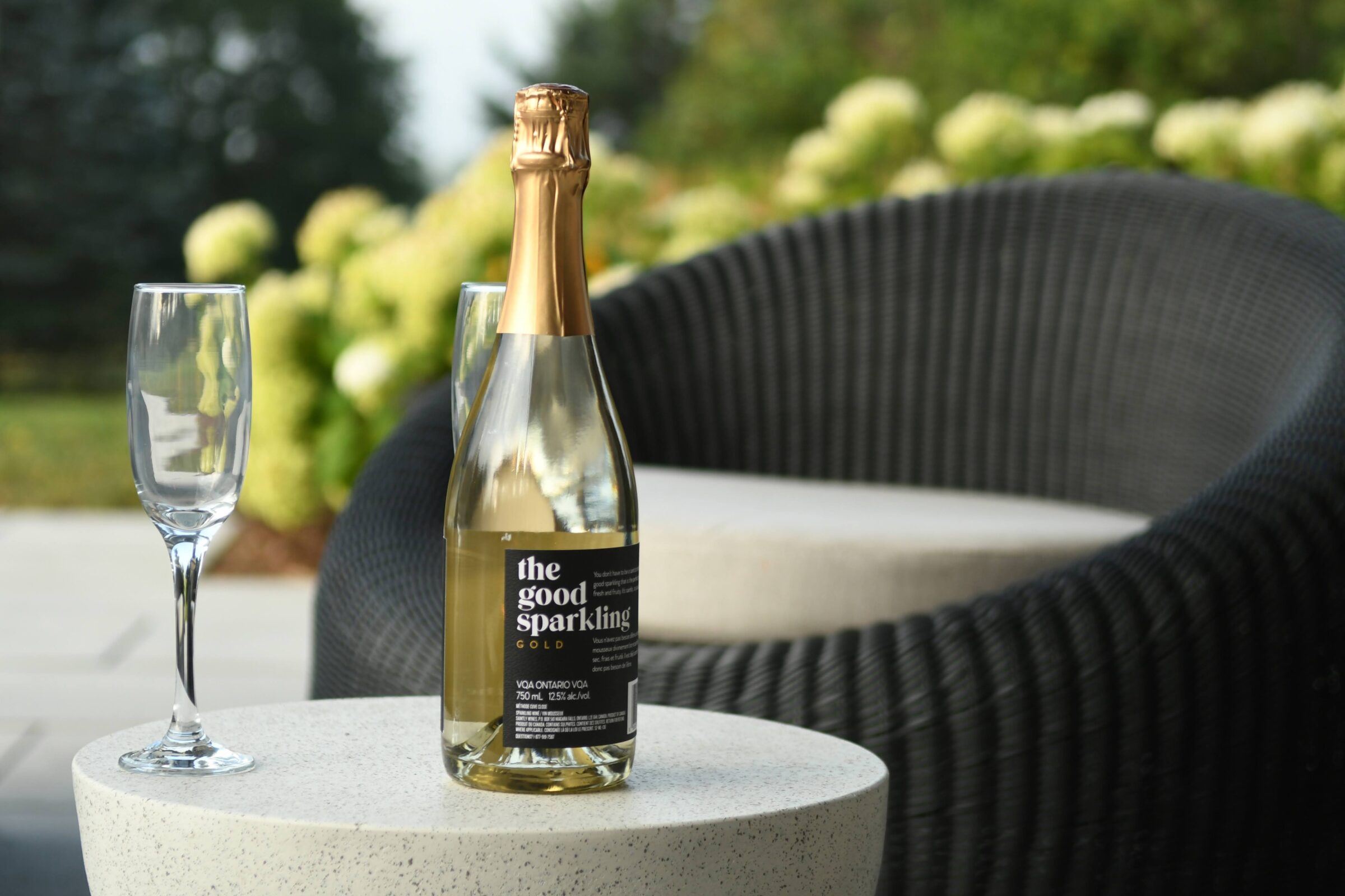A bottle of sparkling wine and a glass on a round table beside a wicker chair, outdoors with greenery in the background.
