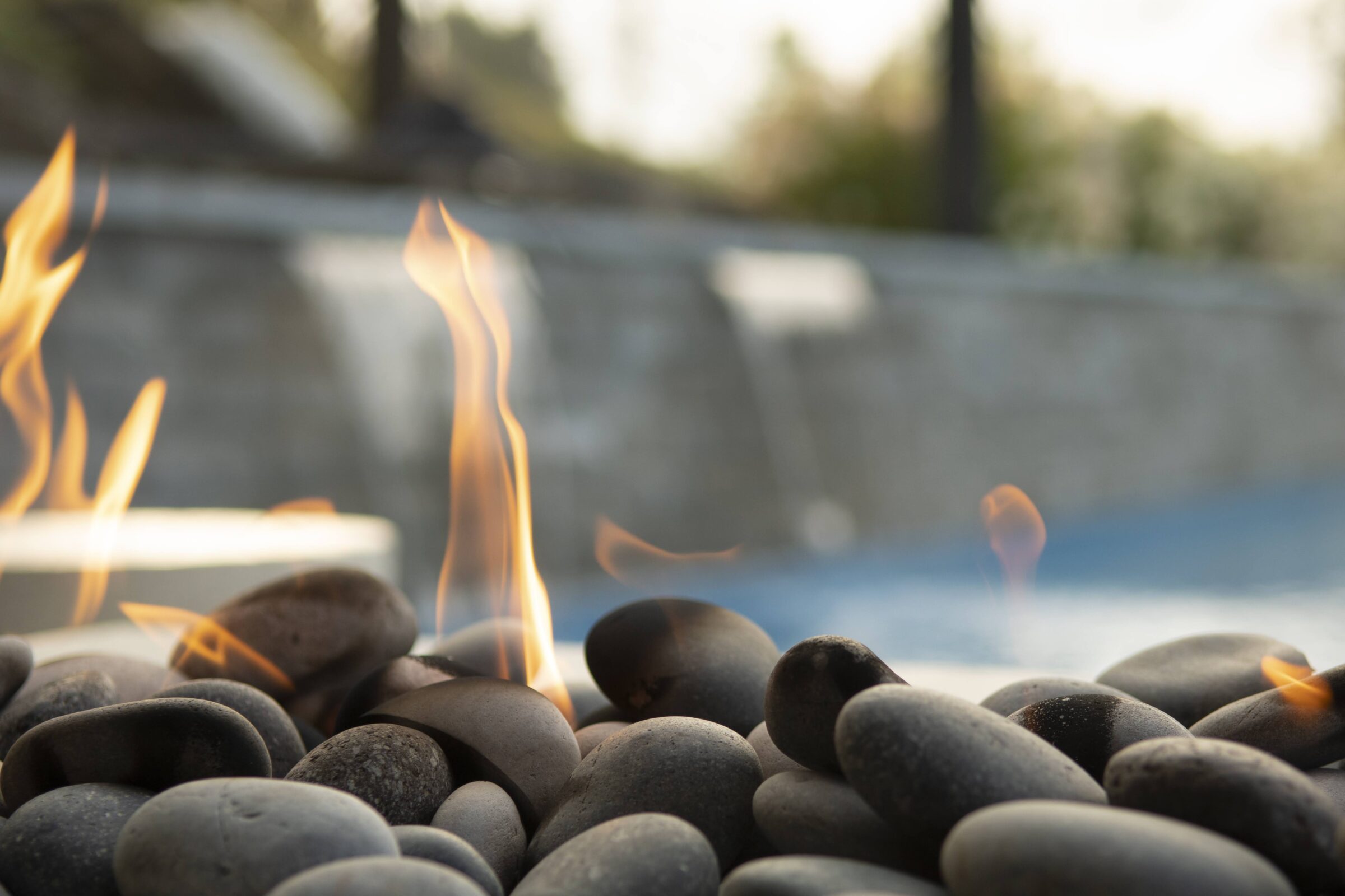Smooth stones with visible flames create a serene, warm ambiance in an outdoor setting, with blurred greenery and stone background.