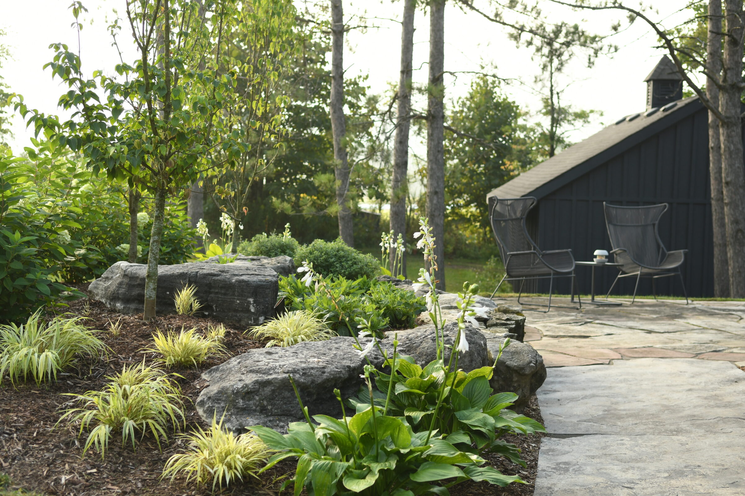 A serene garden scene with lush greenery, stone path, and a black structure. Two chairs and a table sit nearby in tranquility.