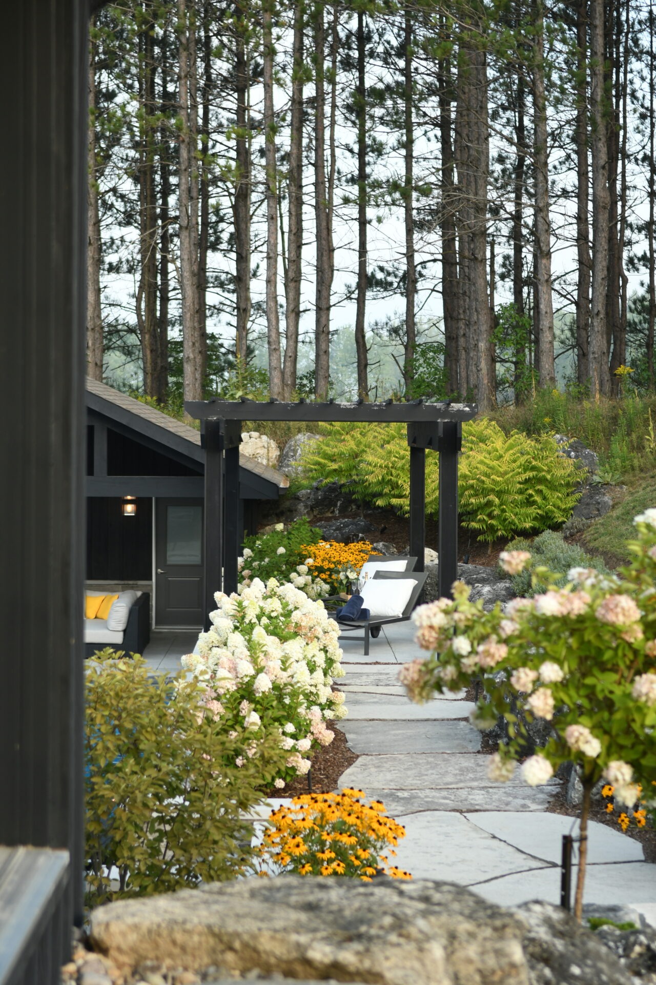 Tranquil garden scene with patio furniture, vibrant flowers, and tall trees. Cozy outdoor seating area next to a modern structure.