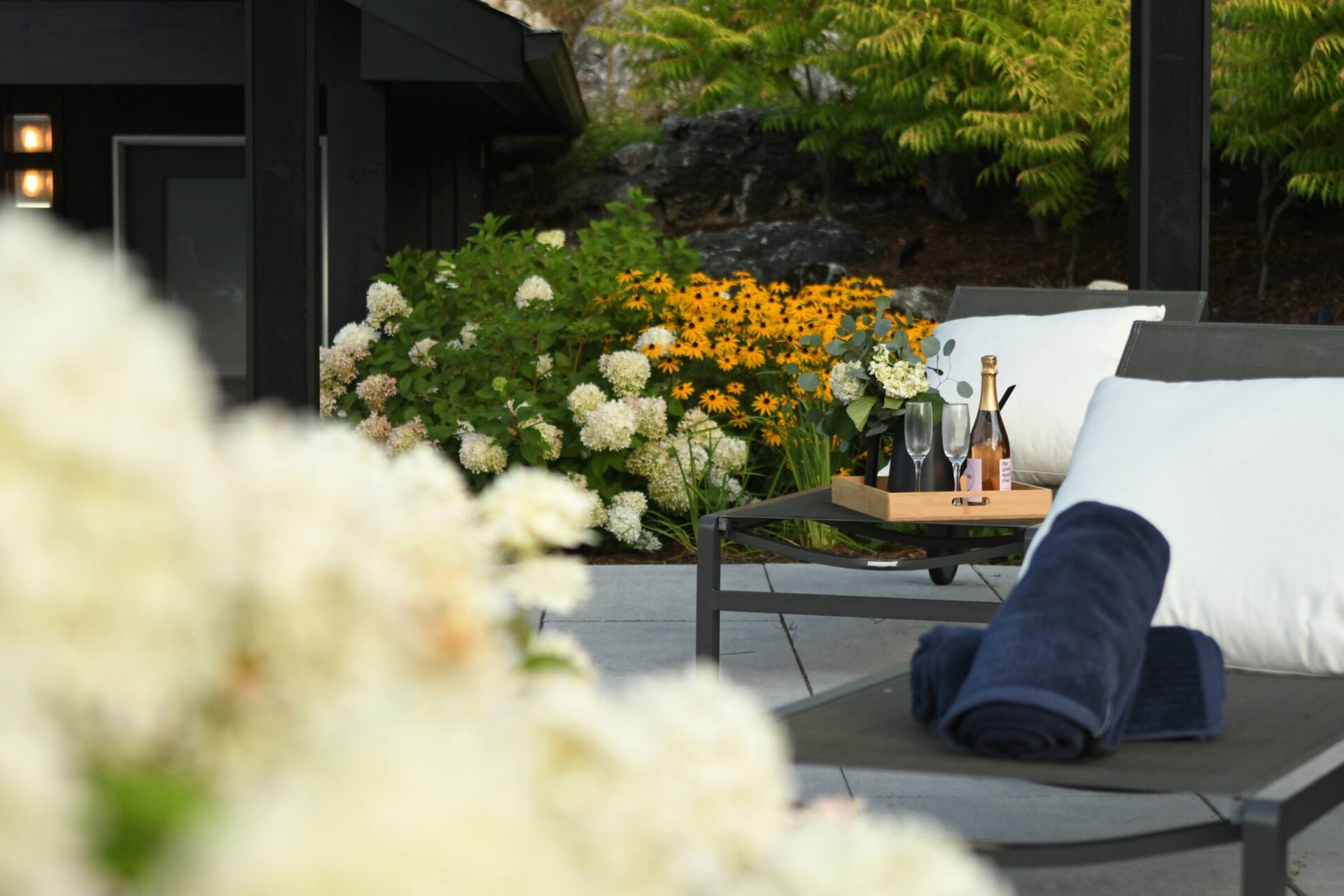 Outdoor lounge area with cushioned chairs, folded towels, table with champagne and glasses, surrounded by lush greenery and blooming flowers. Peaceful ambiance.