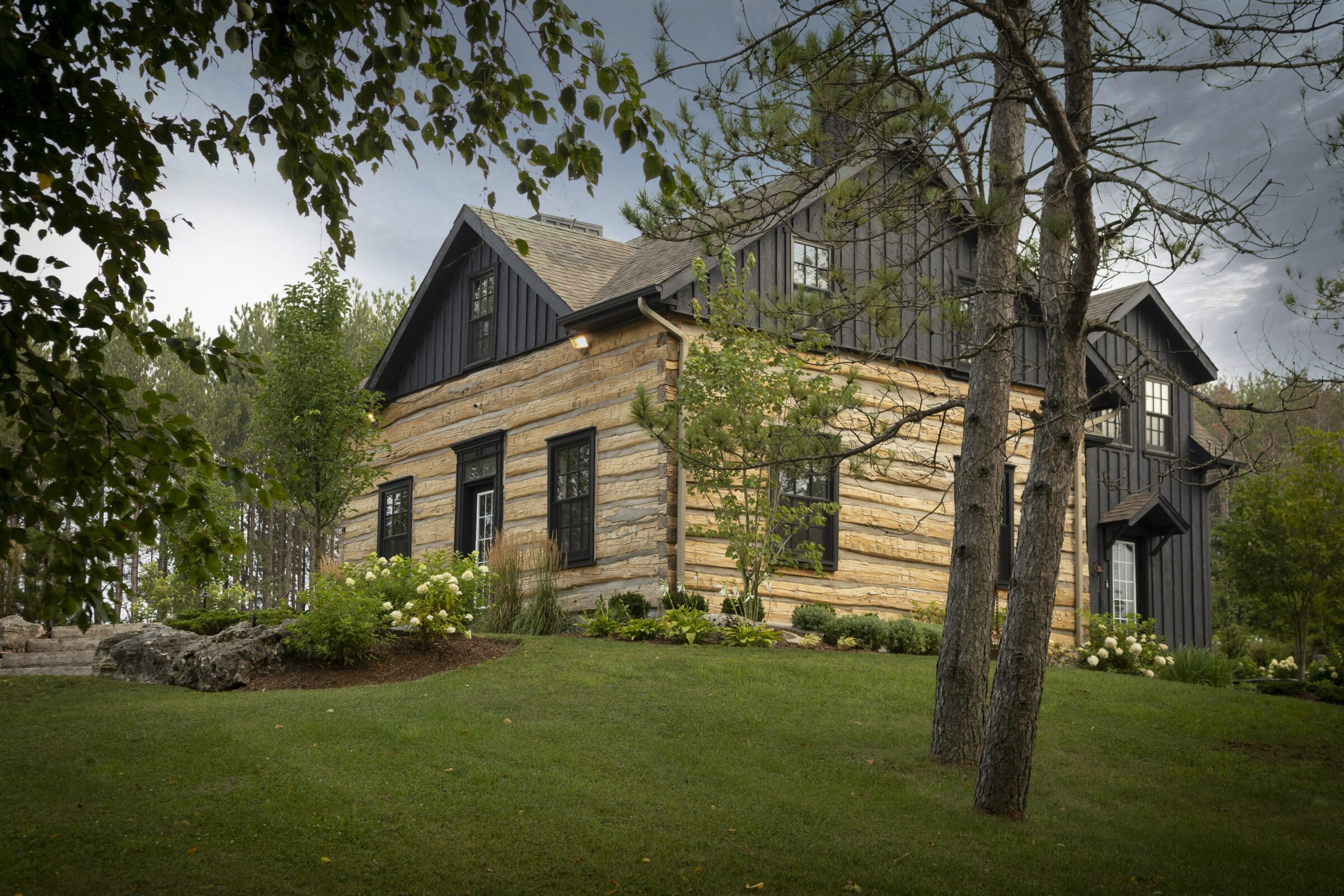 A rustic log cabin with black trim, nestled in lush greenery and trees, under a cloudy sky, conveying a serene countryside ambiance.