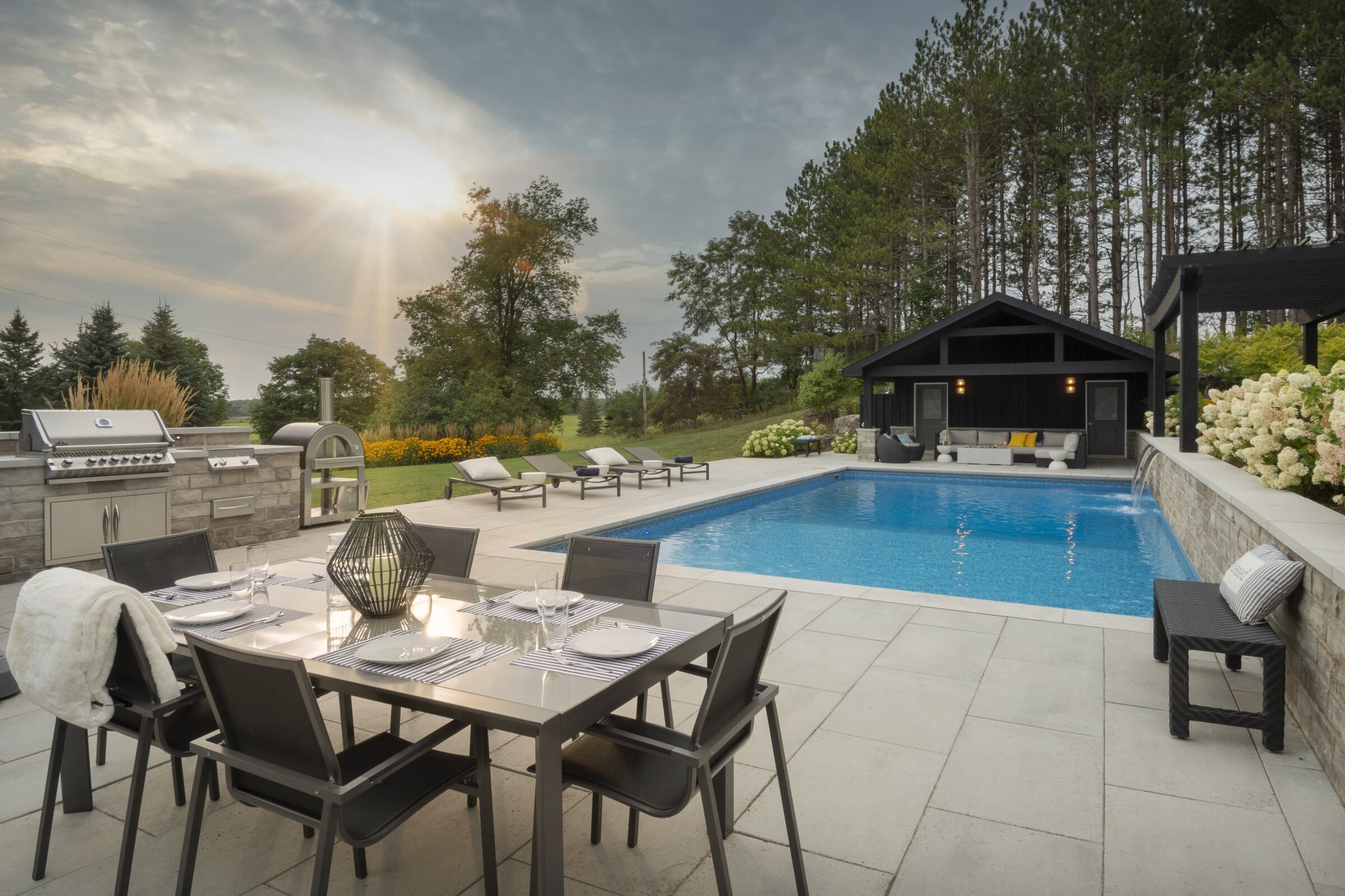 Modern backyard with patio, dining area, outdoor kitchen, and pool. Surrounded by trees, open sky, and landscaped gardens, creating a serene setting.