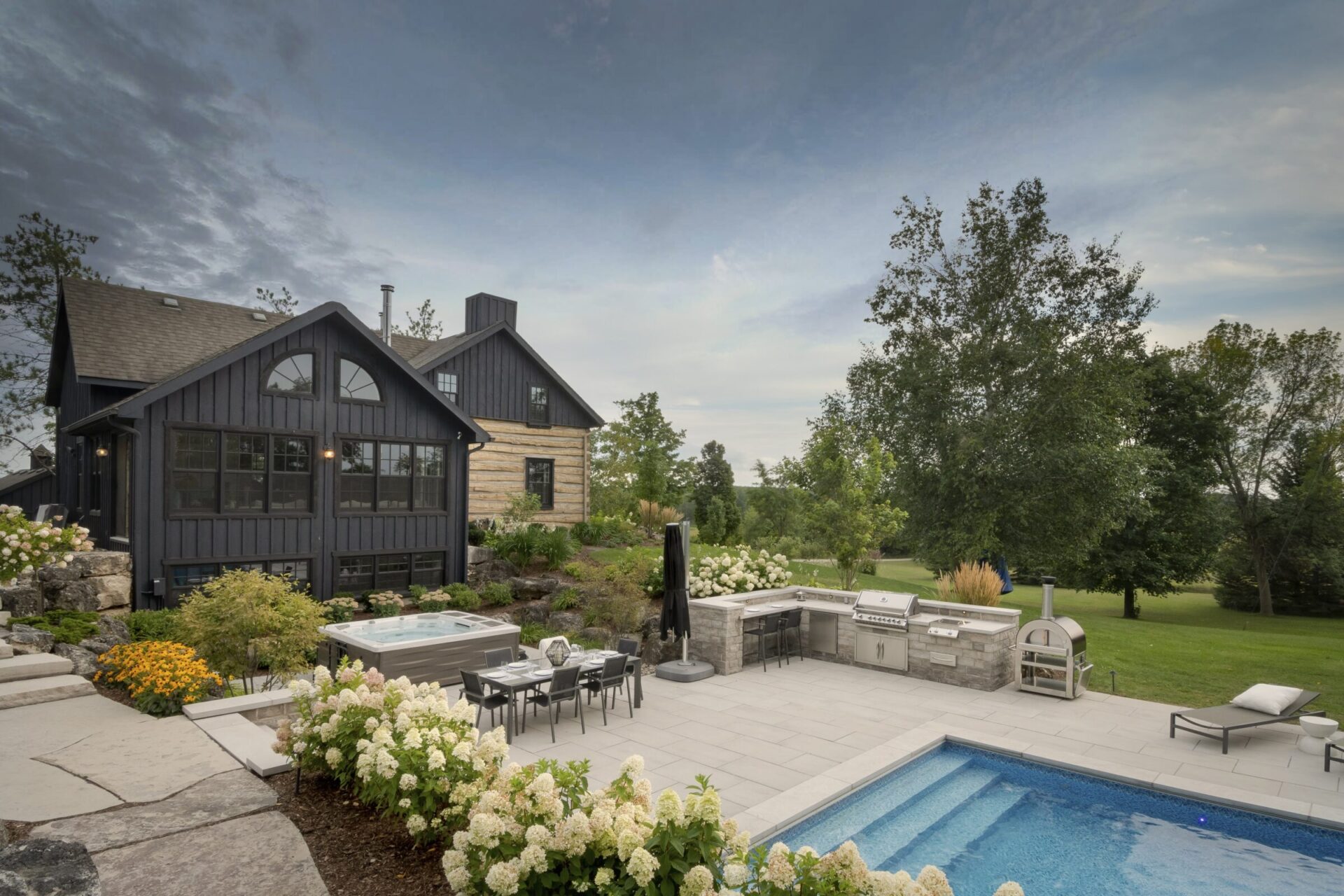 Modern backyard with hot tub, dining area, and outdoor kitchen beside a cozy house. Surrounded by lush greenery and blooming flowers.