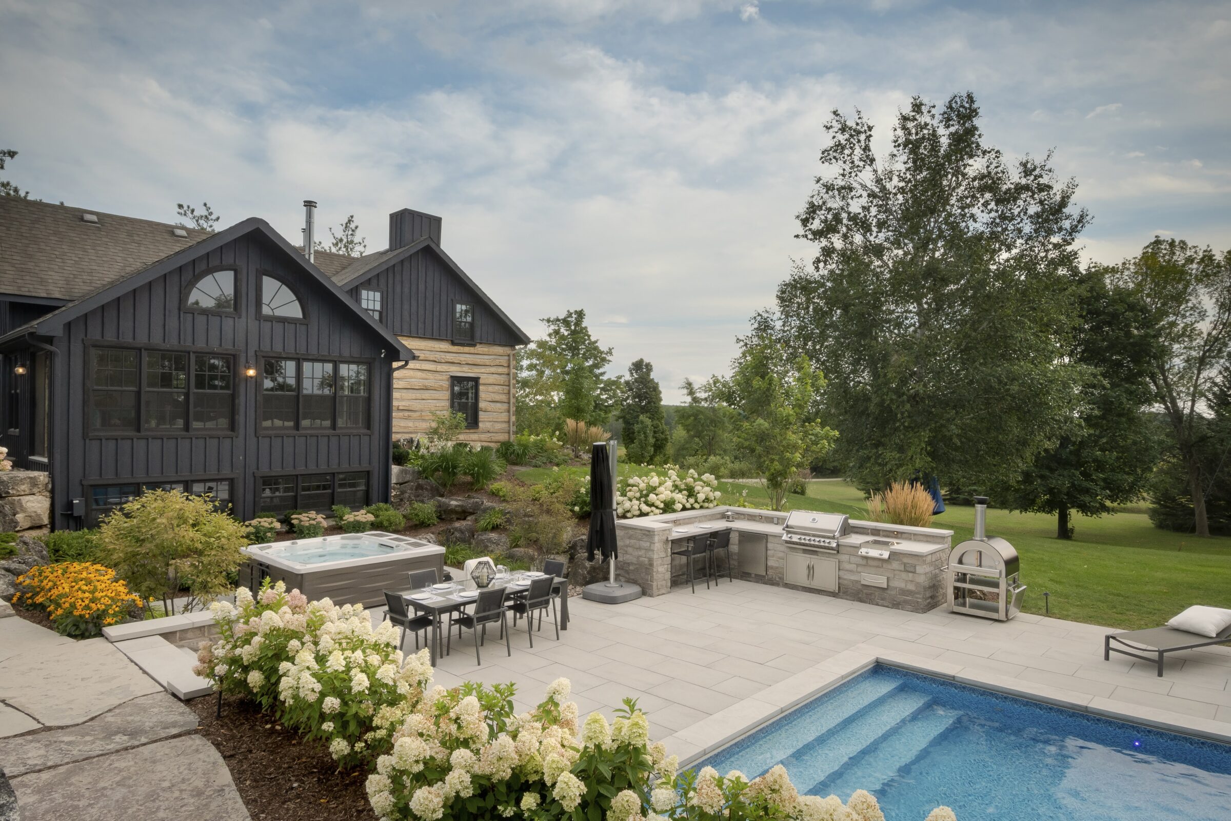 A modern countryside home features a stone patio, swimming pool, and outdoor kitchen surrounded by lush greenery and blooming white flowers.