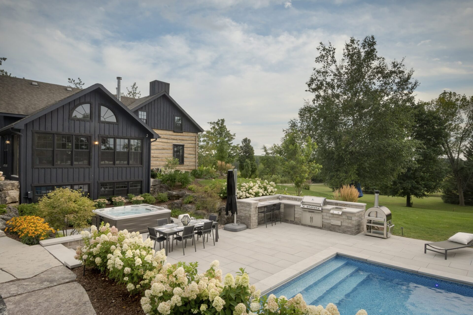 A modern house with a porch overlooks a backyard featuring a pool, patio, outdoor kitchen, and lush garden landscaping under a cloudy sky.