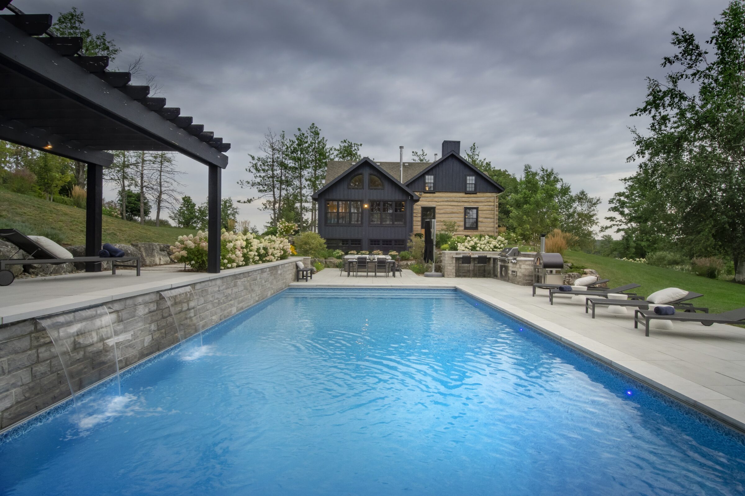 A modern home with a swimming pool, lounge chairs, pergola, and lush greenery under a cloudy sky, creates a serene outdoor setting.