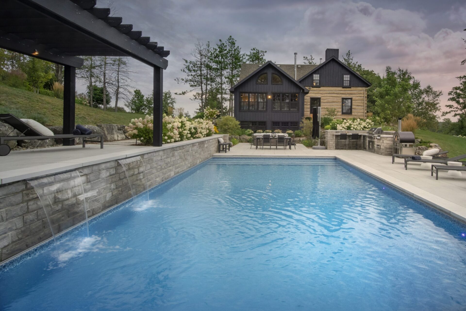 A serene backyard with a swimming pool, stone patio, pergola, barbecue area, surrounded by greenery and a charming black house.