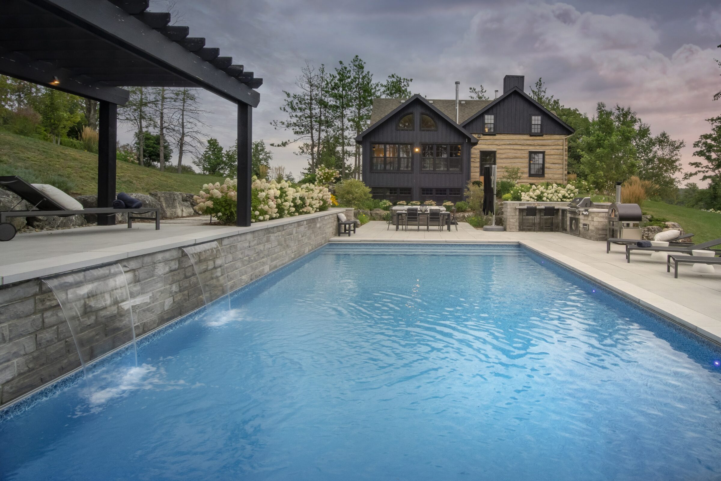 A serene backyard with a swimming pool, stone patio, pergola, barbecue area, surrounded by greenery and a charming black house.