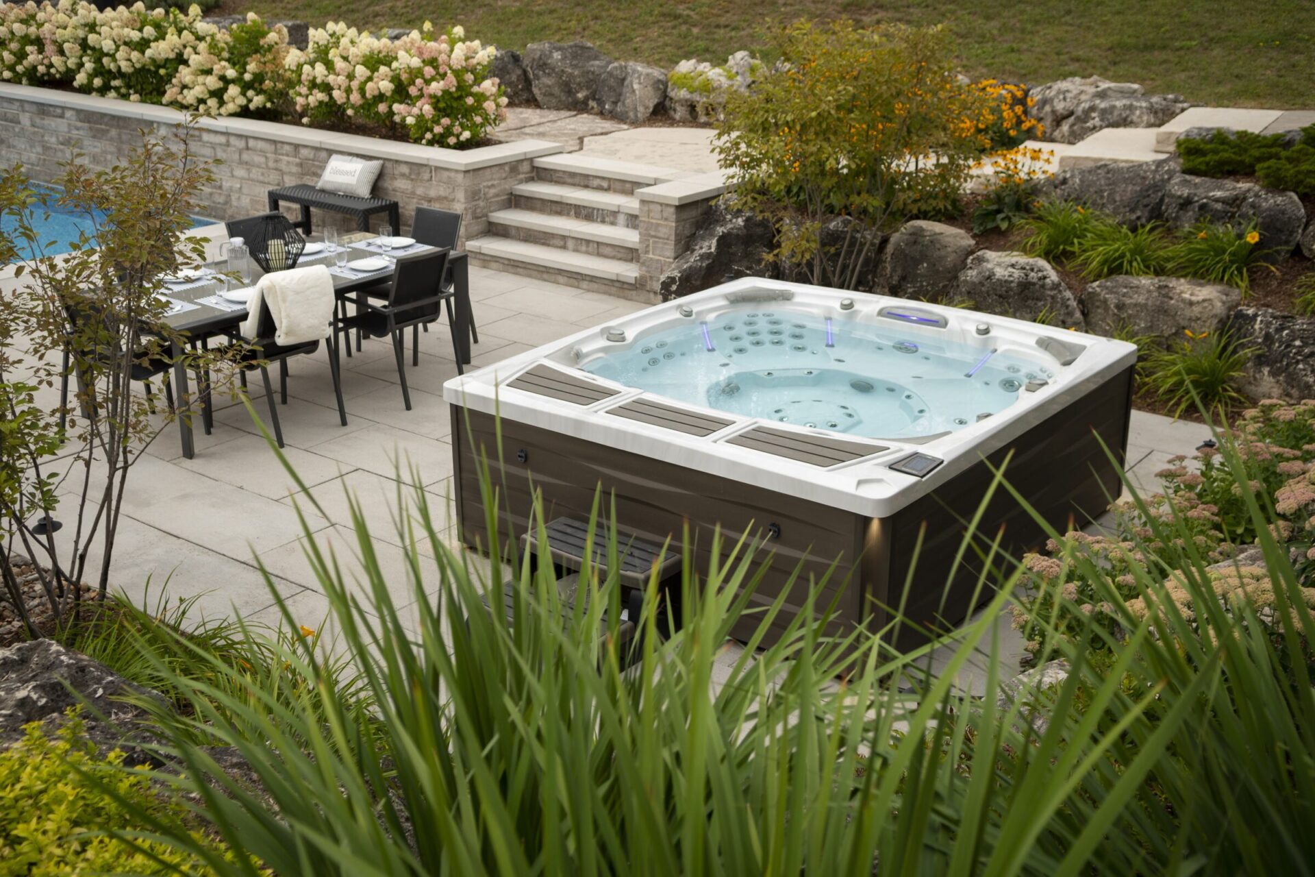 A luxurious backyard scene with a hot tub, neatly set dining table, surrounded by stone landscaping, flowers, and greenery. Peaceful and inviting ambiance.