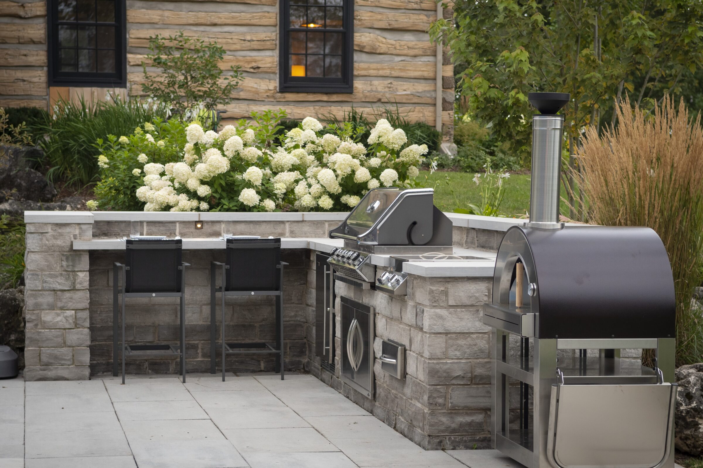 Outdoor kitchen with stone counters, bar stools, grill, and pizza oven. Surrounded by lush greenery and a wooden house backdrop.