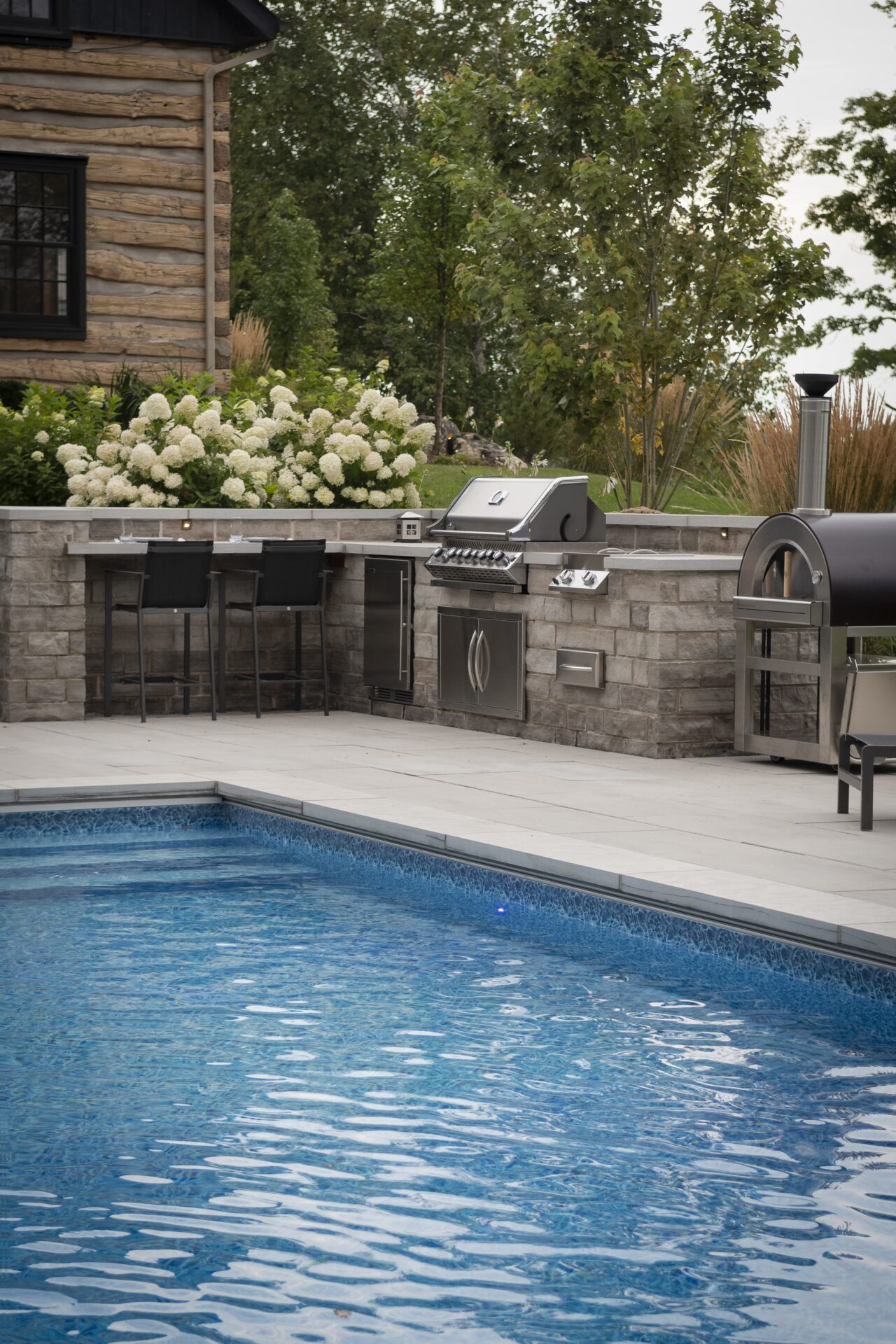 A modern patio with a pool, outdoor kitchen, and grill near a log cabin, surrounded by lush greenery and flower bushes.