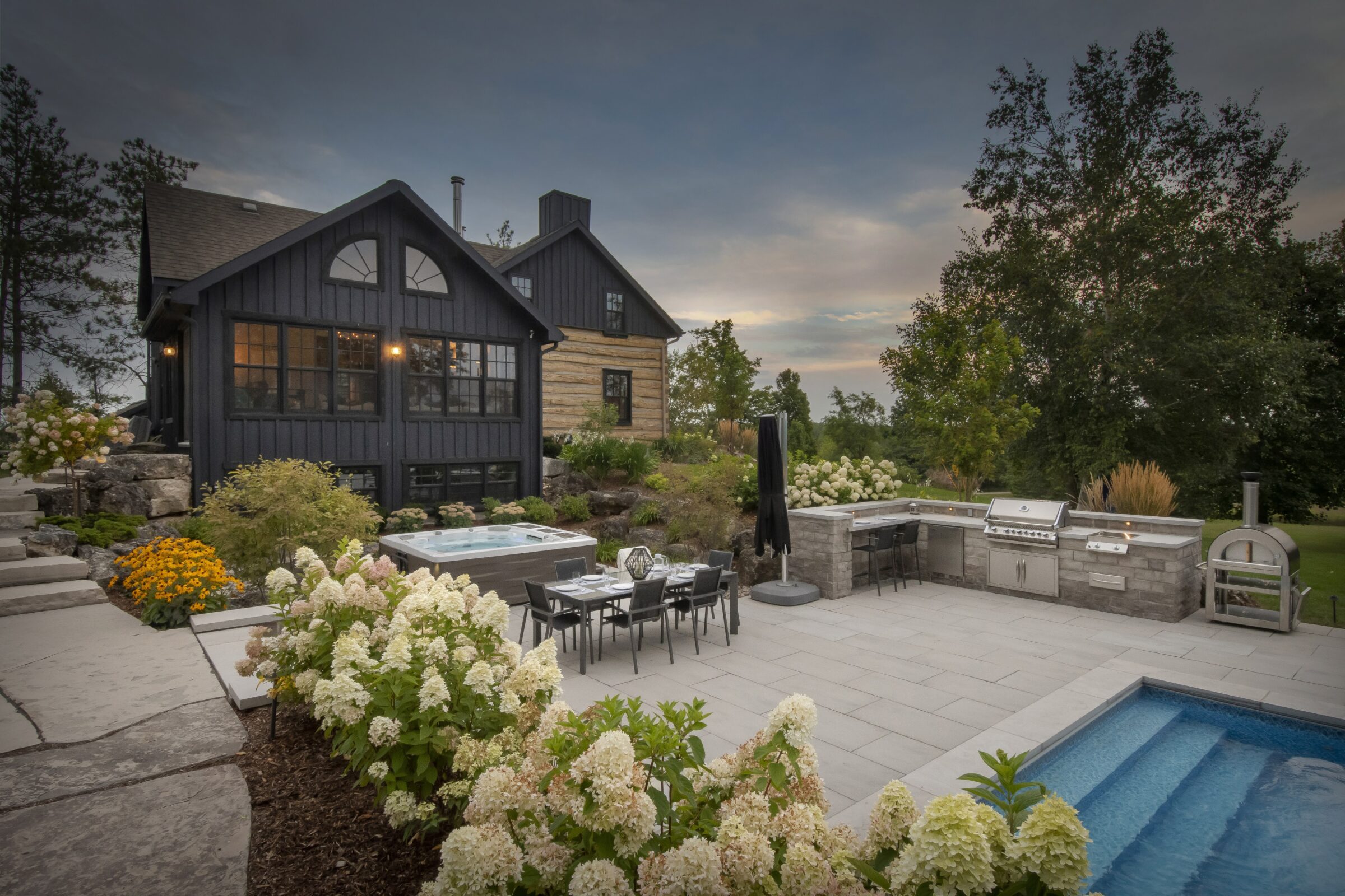 A cozy modern house with lush garden, outdoor kitchen, dining area, jacuzzi, and pool, surrounded by trees and blooming flowers under evening sky.