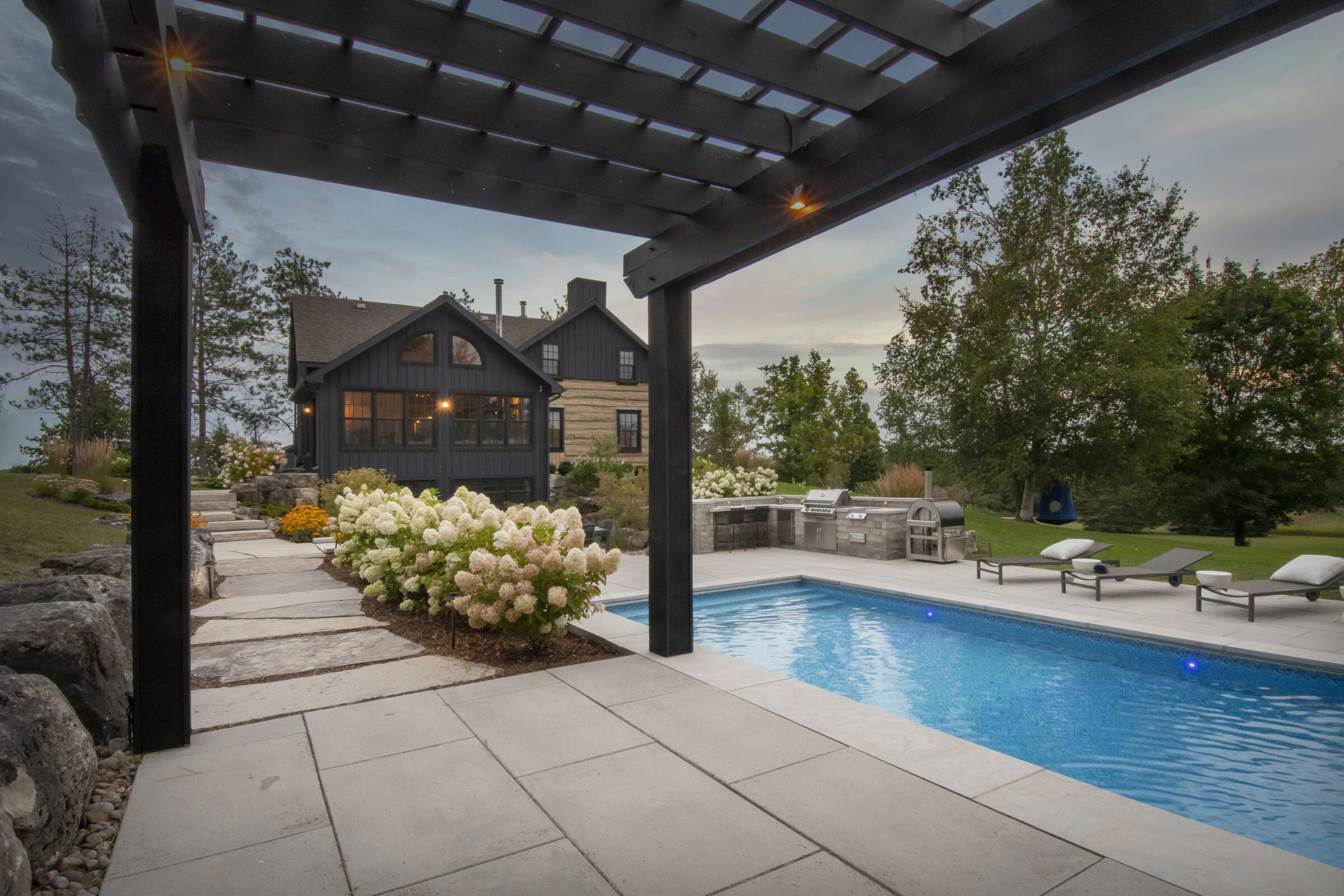 A modern house with a wooden pergola overlooks a pool, surrounded by landscaped gardens and trees, with outdoor grill area and loungers.