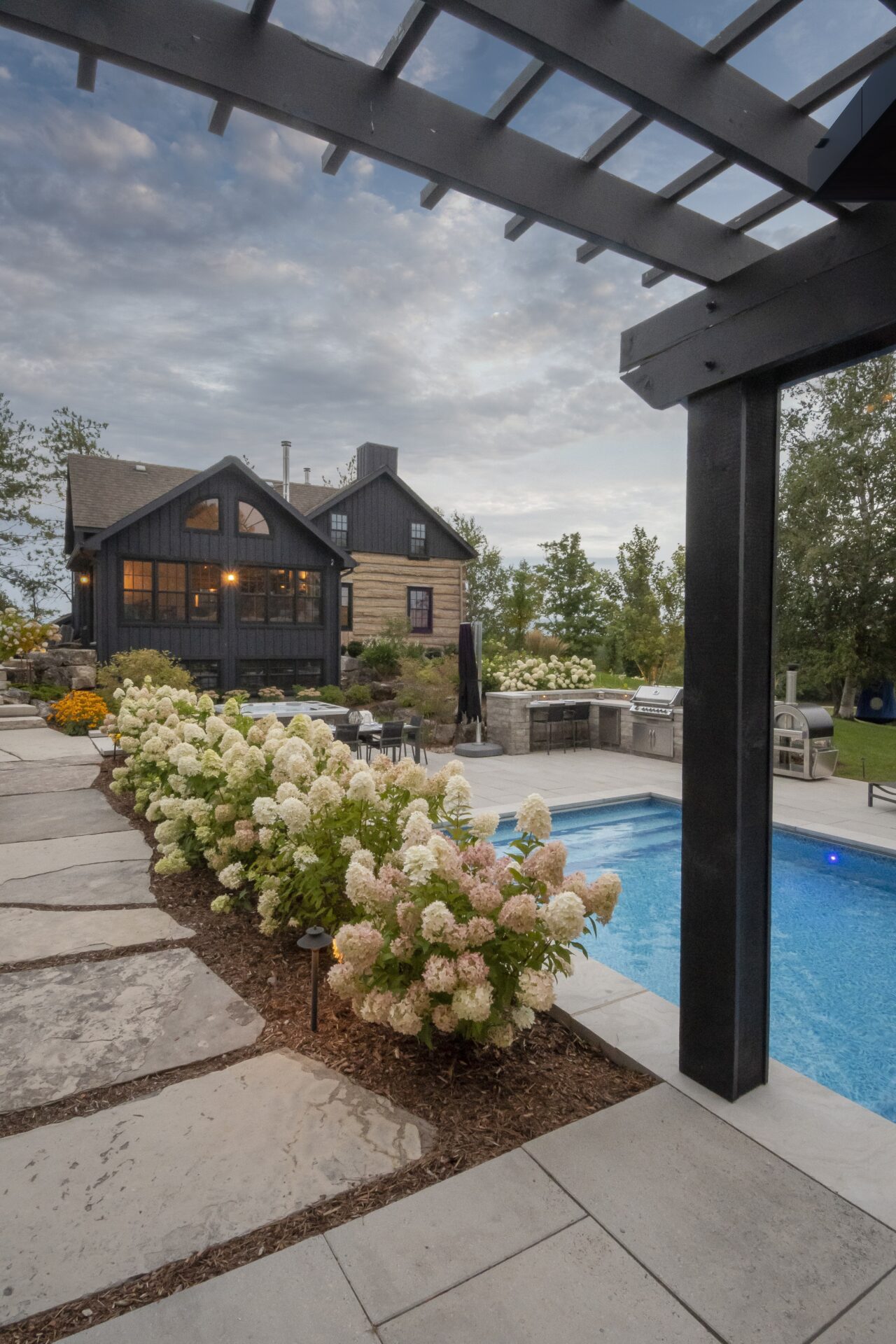 Charming house with wooden exterior, swimming pool, and patio. Lush floral landscaping lines the stone pathway, creating a serene backyard oasis under cloudy skies.