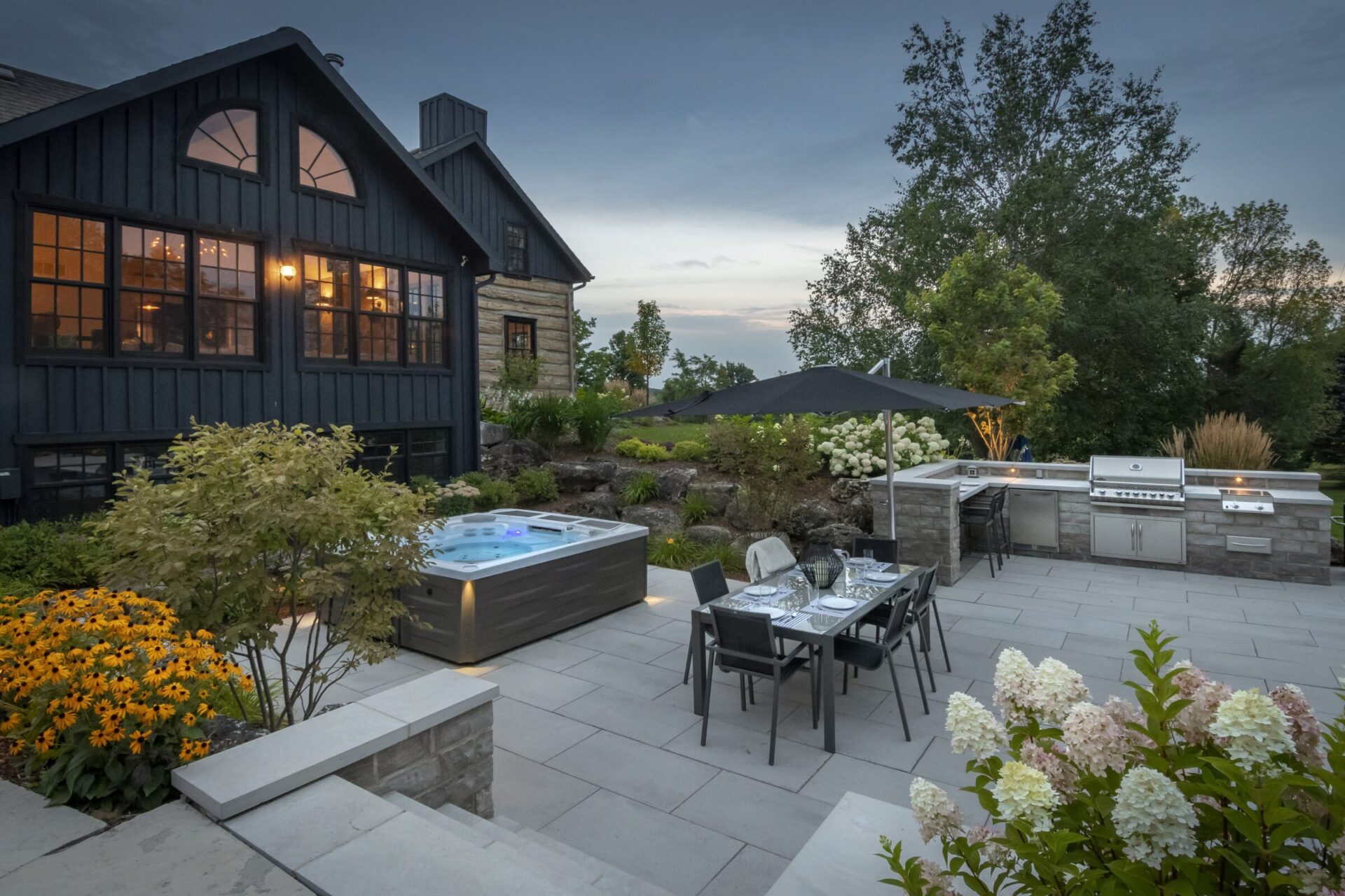 Modern outdoor patio with dining table, hot tub, and barbecue area surrounded by lush landscaping and a cozy house with glowing windows.