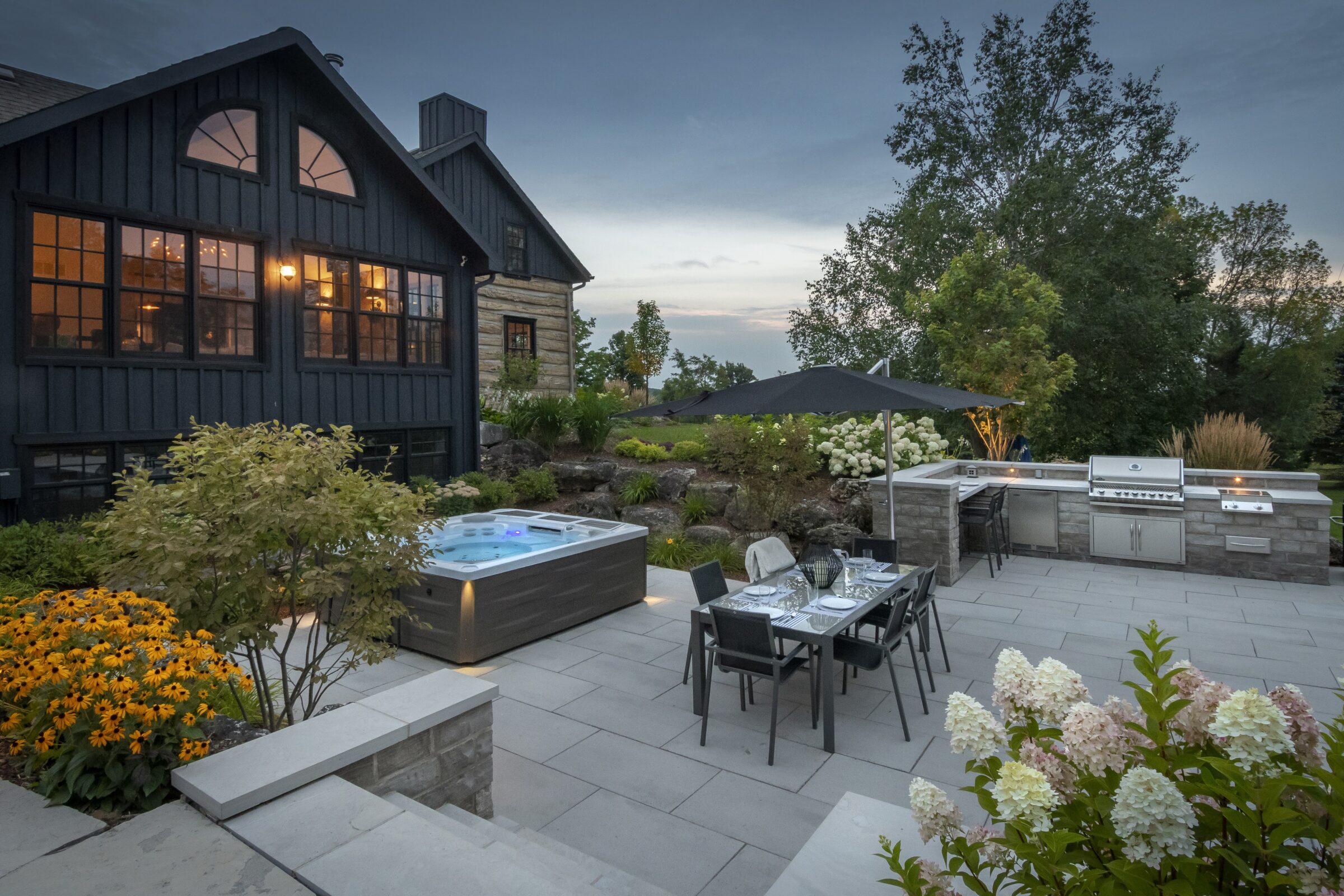 Modern patio with hot tub, dining set, outdoor kitchen, and garden. Dark house backdrop. Tranquil evening setting with trees and flowers.