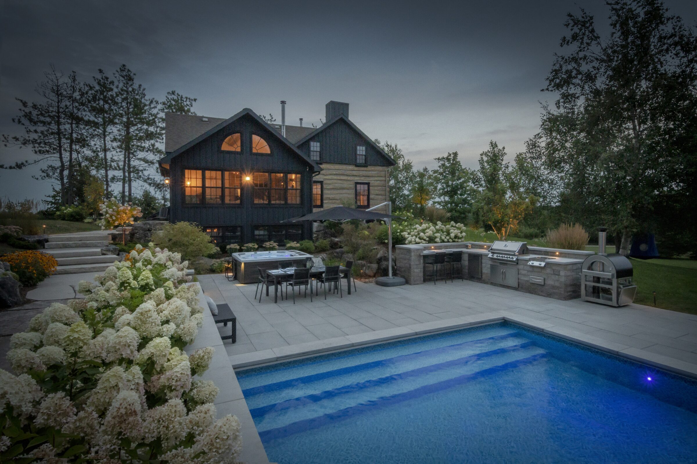 A cozy two-story house with a lit patio, swimming pool, and outdoor kitchen, surrounded by landscaped gardens and trees in the evening light.