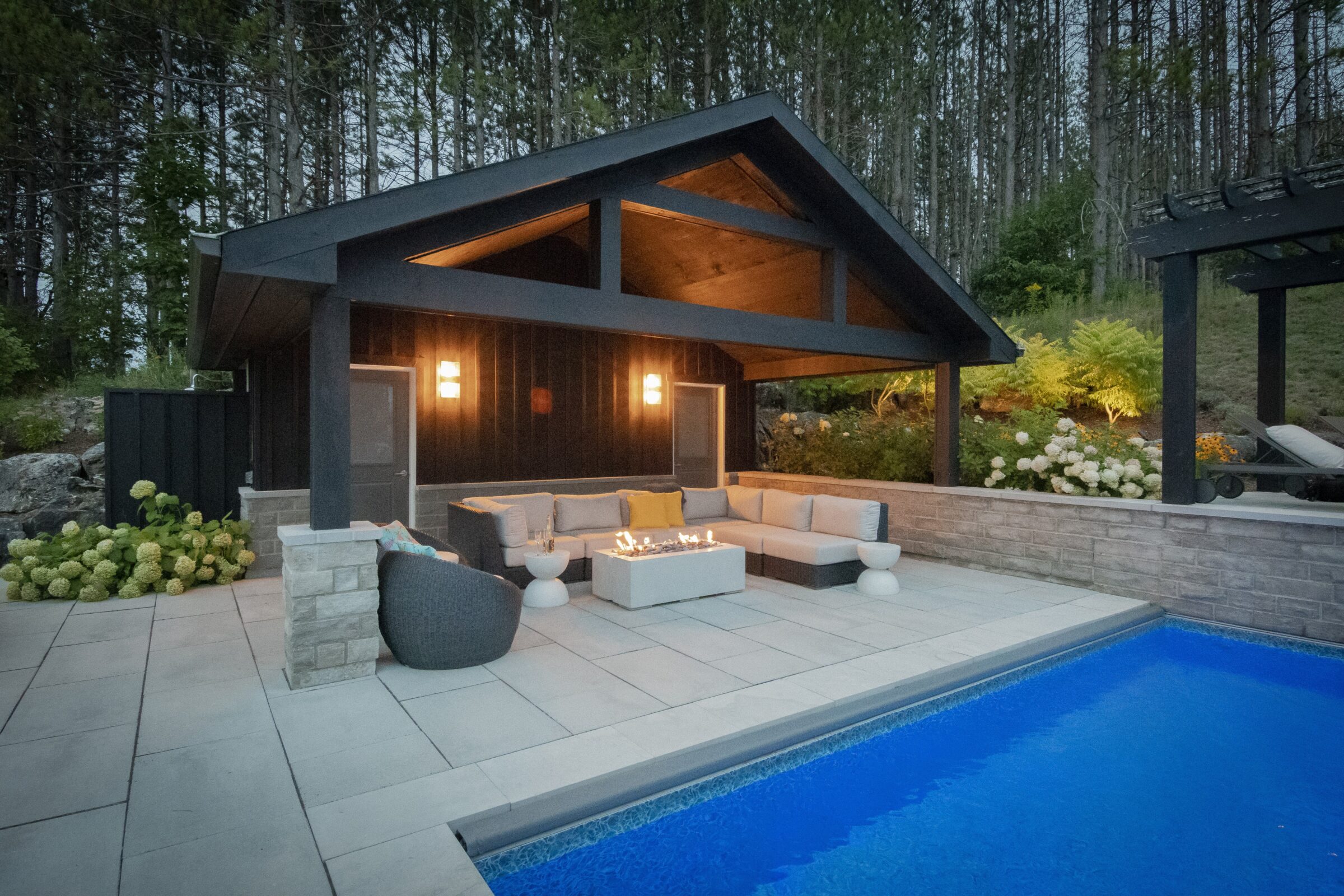 Cozy outdoor lounge with modern fire pit, pool, and natural surroundings. Illuminated seating area sheltered by a stylish canopy amid lush greenery.