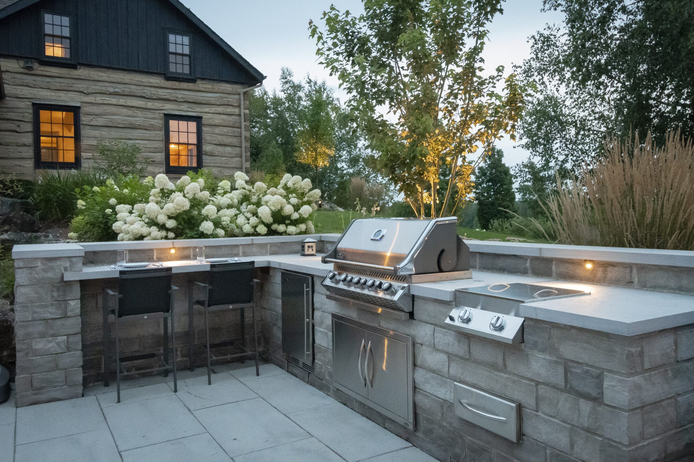 Outdoor kitchen with modern grill, bar seating, and counters. Illuminated patio with greenery and flowers. Rustic log house in the background.