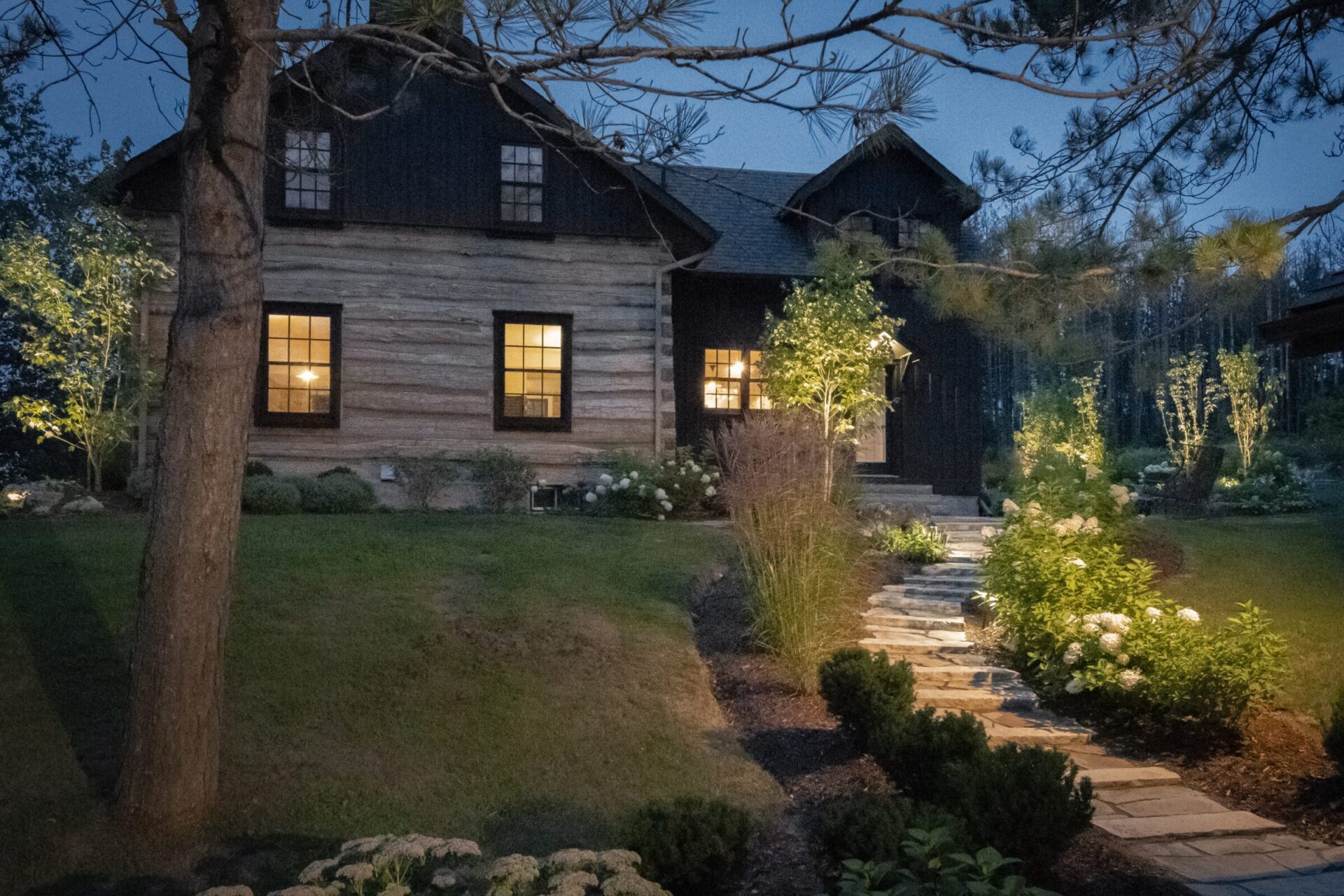 A cozy, rustic cabin with glowing windows and a stone path, surrounded by lush greenery and softly lit at dusk.