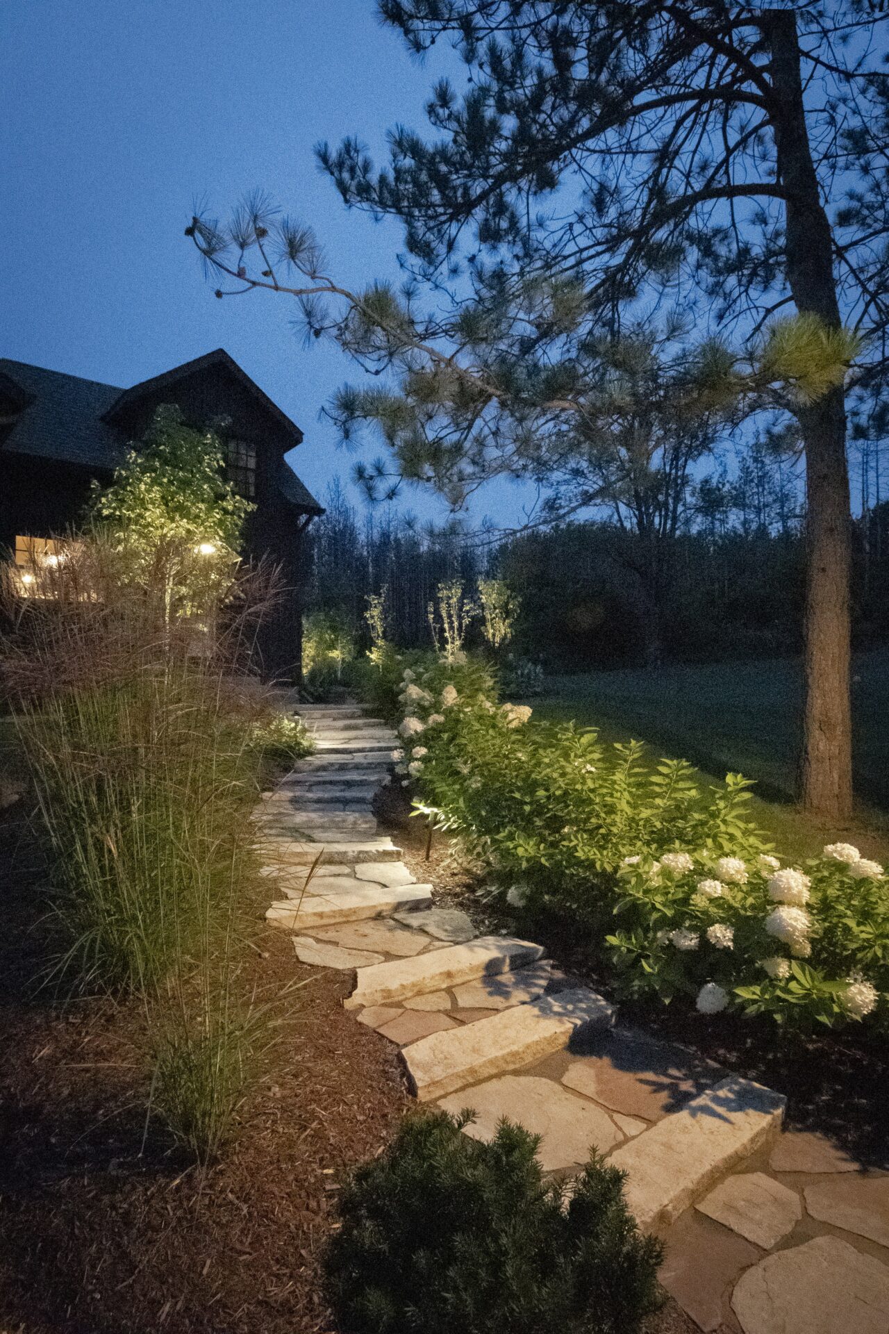 A stone path leads to a house with glowing windows, surrounded by trees and flowering shrubs, under a twilight sky.