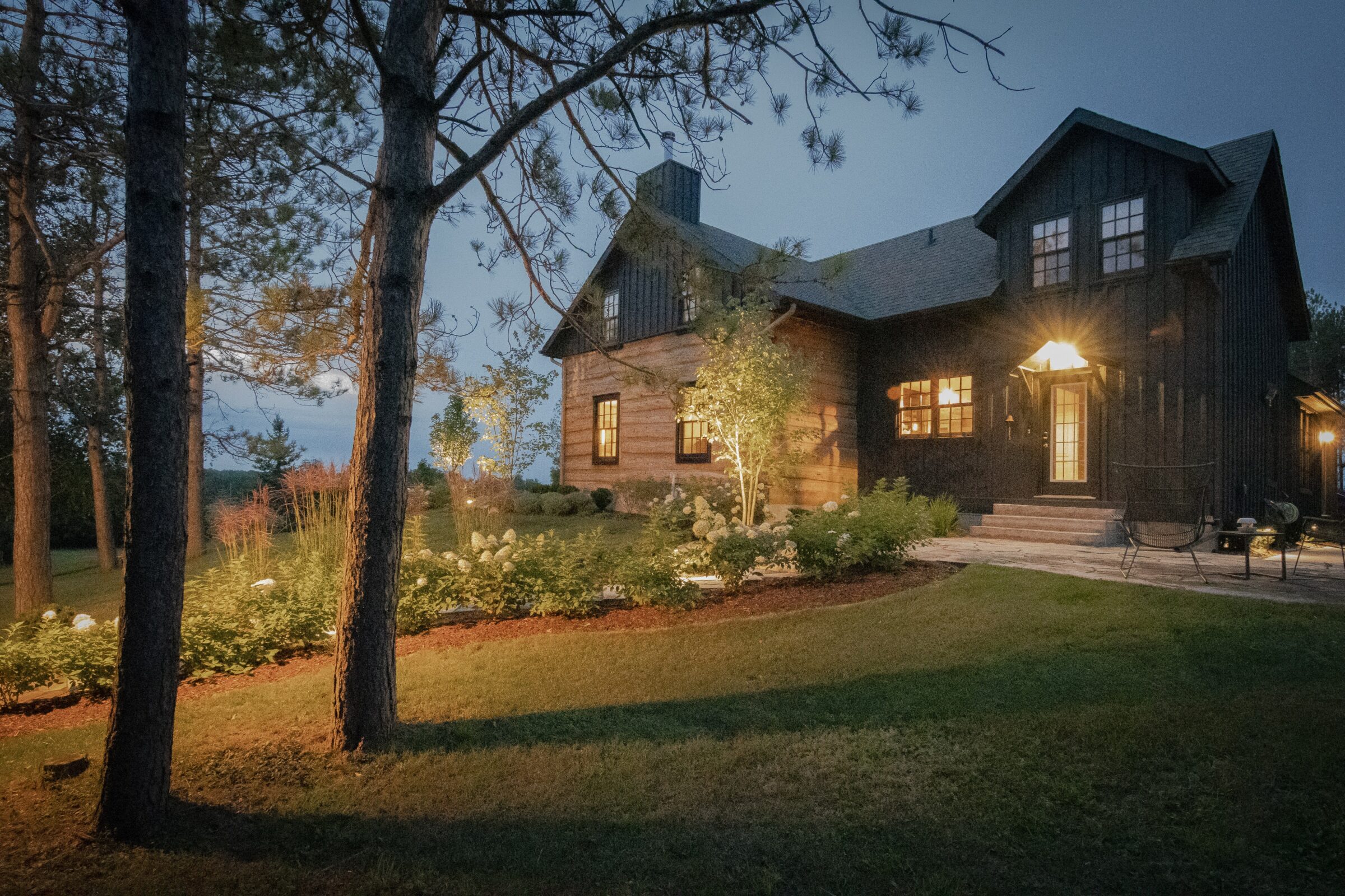 Cozy wooden house at dusk, surrounded by trees and well-manicured garden, warmly lit windows enhancing the peaceful, inviting atmosphere.